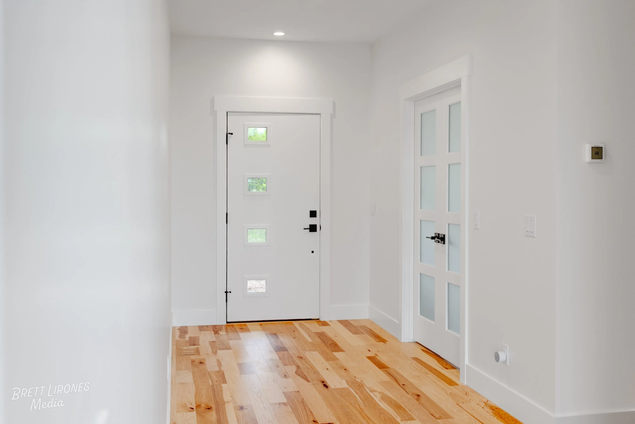Empty entryway with white walls, light hardwood floors, a white front door with four small square windows, and a white interior door with frosted glass panels.