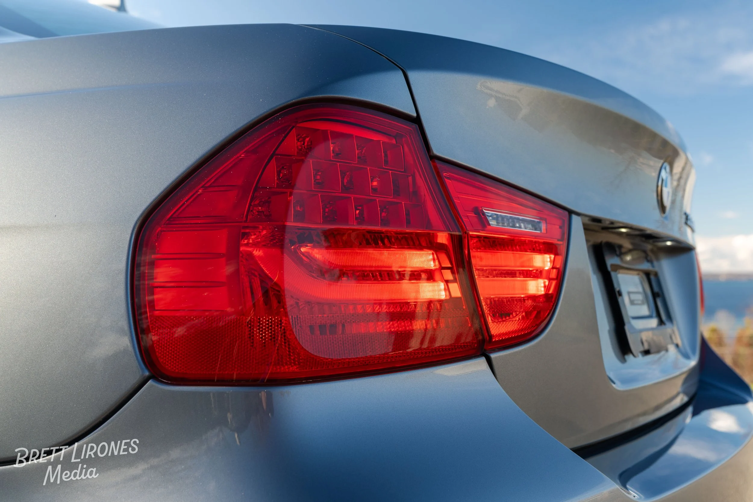 Close-up of the rear red tail light of a silver BMW car with a blue sky background.