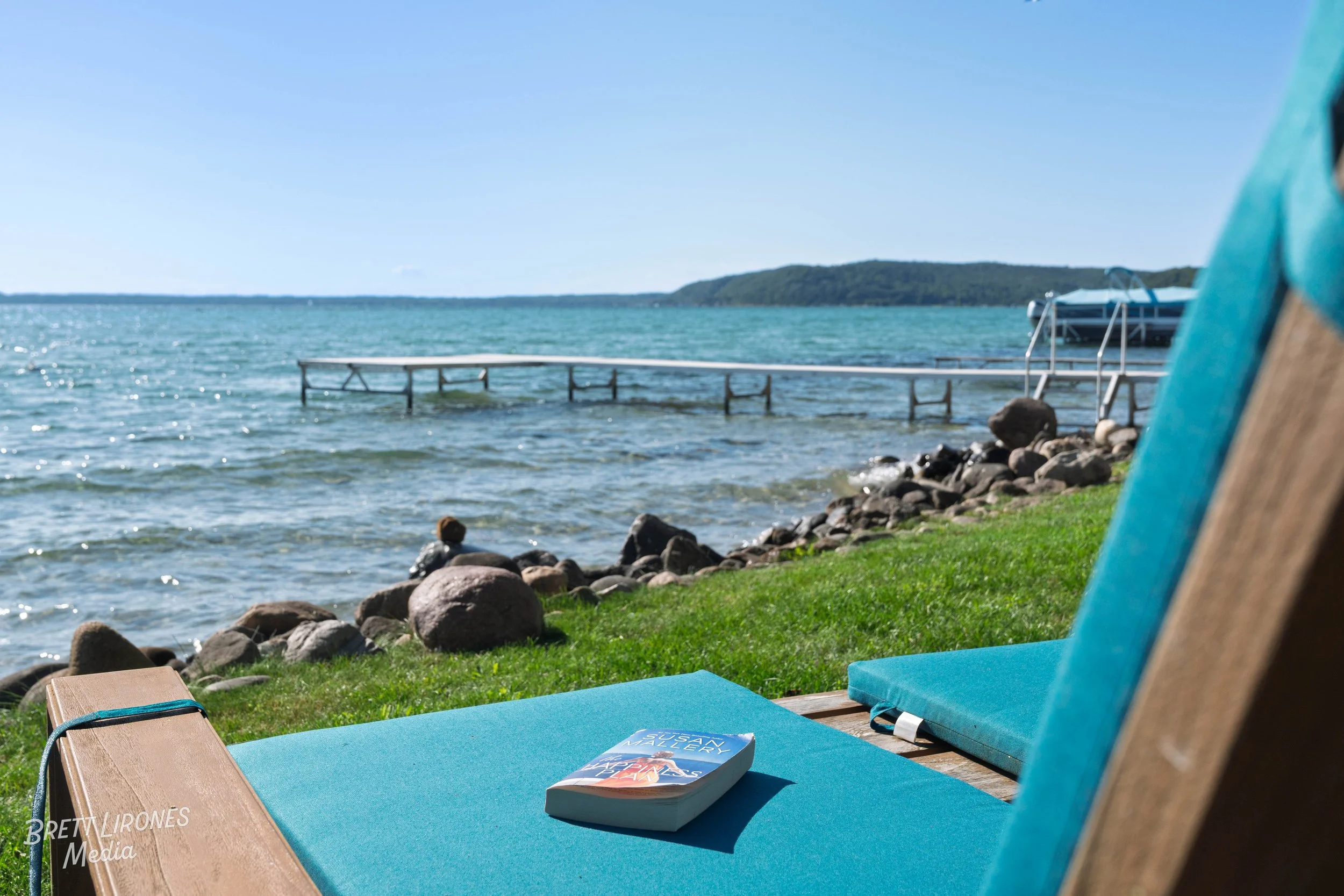 A lakeside scene on a sunny day with a wooden lounge chair with blue cushions and a book on it, near a grassy area with rocks, a dock extending into the water, and a boat in the background.