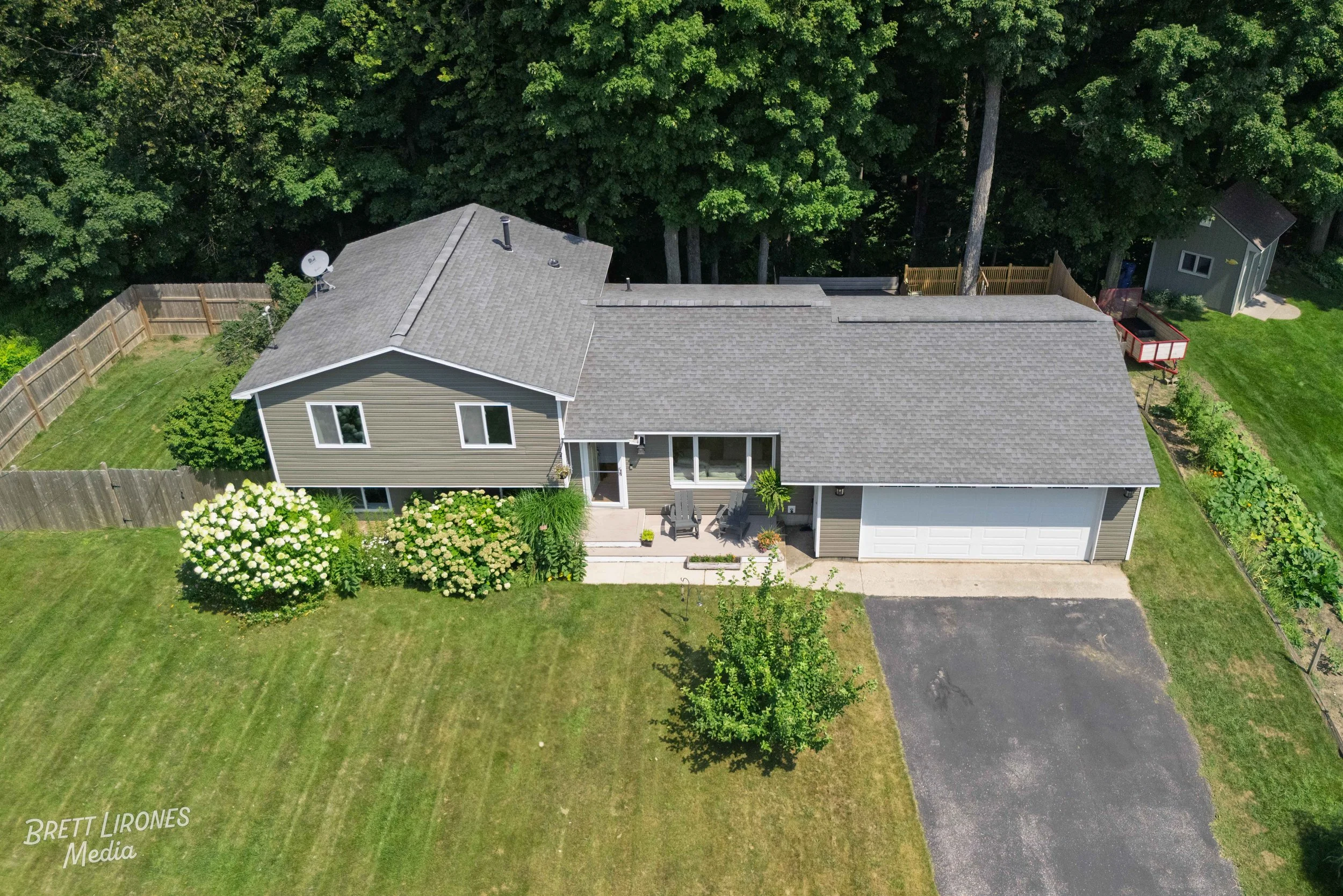 Aerial view of a suburban house with a gray roof, beige siding, a white garage door, a small front porch with chairs, surrounded by a lush lawn, trees, and garden beds.