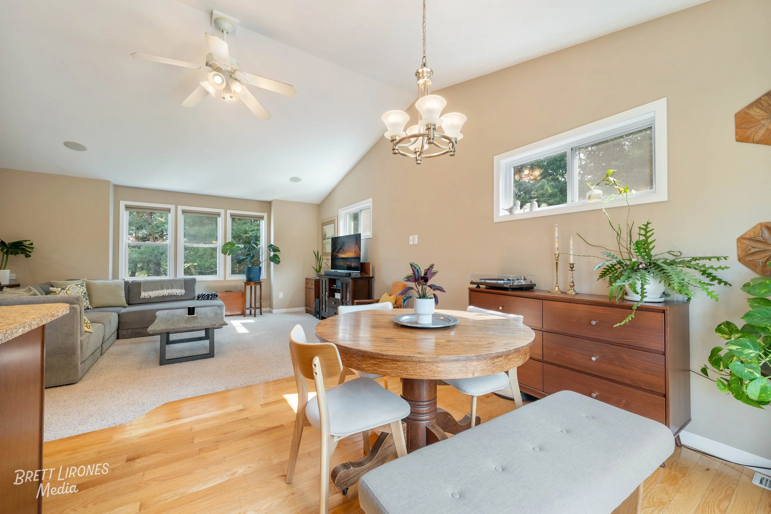 Living room with a gray sectional sofa, a wooden dining table with chairs, a wooden dresser, and multiple windows with greenery outside.