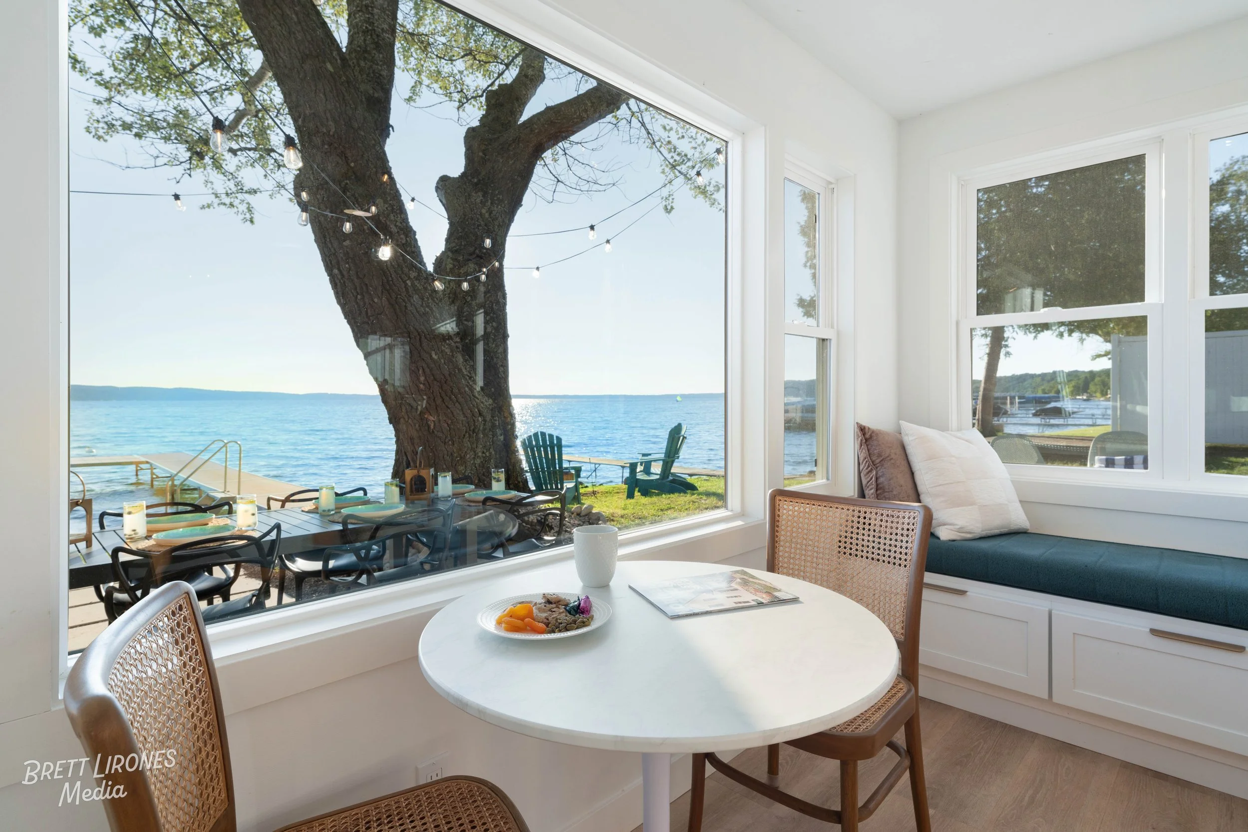 Interior view of a dining nook with a white round table, two wooden chairs, and a window seat with cushions, overlooking a lakeside scene with trees, a dock, Adirondack chairs, and string lights outside.
