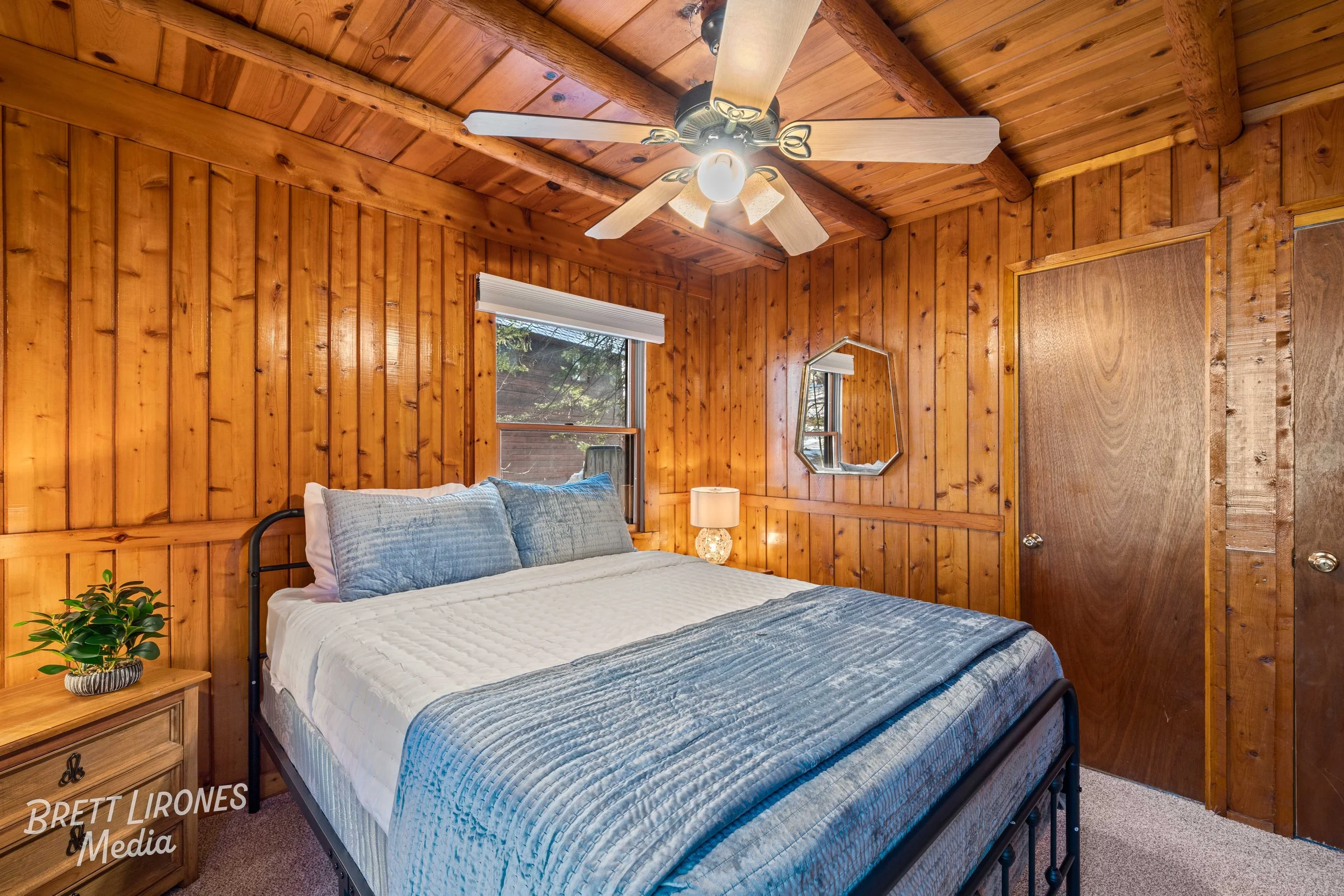 A cozy bedroom with wood-paneled walls and ceiling, a bed with white and blue bedding, a nightstand with a plant, a small lamp, a window with white blinds, and a mirror on the wall.