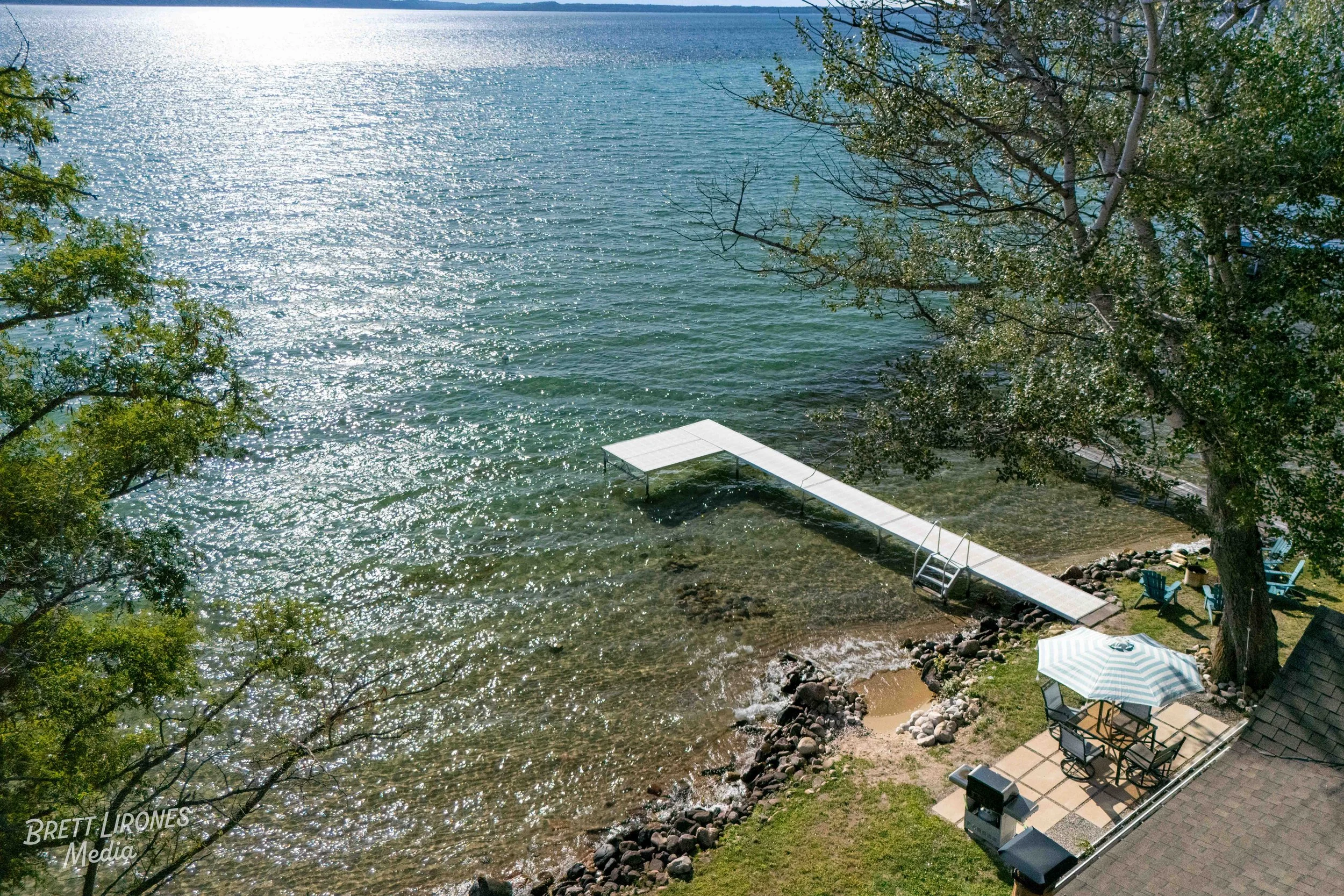 Aerial view of a lakeside backyard with a dock, a large tree, two Adirondack chairs, a table with an umbrella, and a small barbecue grill.