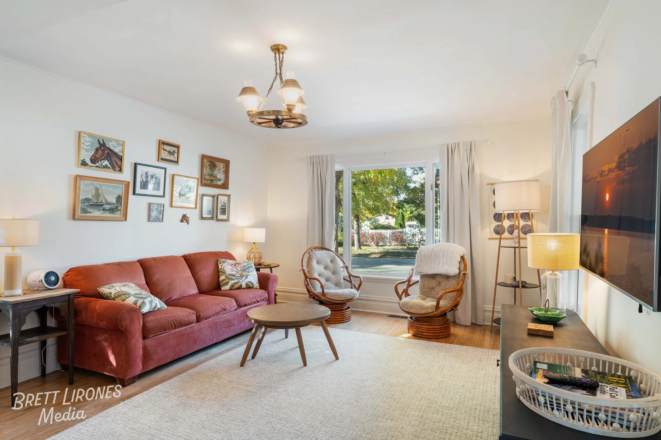 Living room with a red sofa, eclectic wall art, two cushioned wicker chairs by a window, and a large flatscreen TV on a stand, decorated with lamps and a small basket of magazines on top.