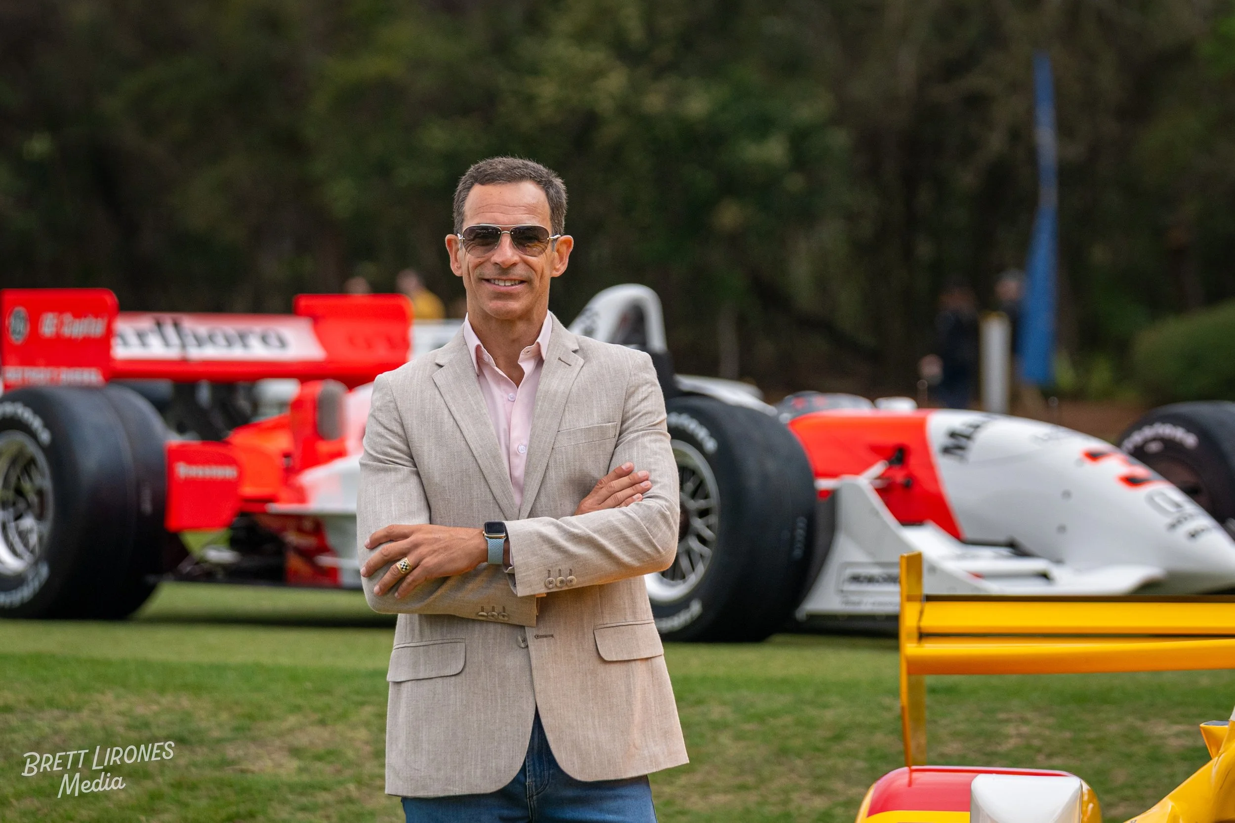 A man in a beige blazer and sunglasses stands with arms crossed in front of a vintage race car outdoors.