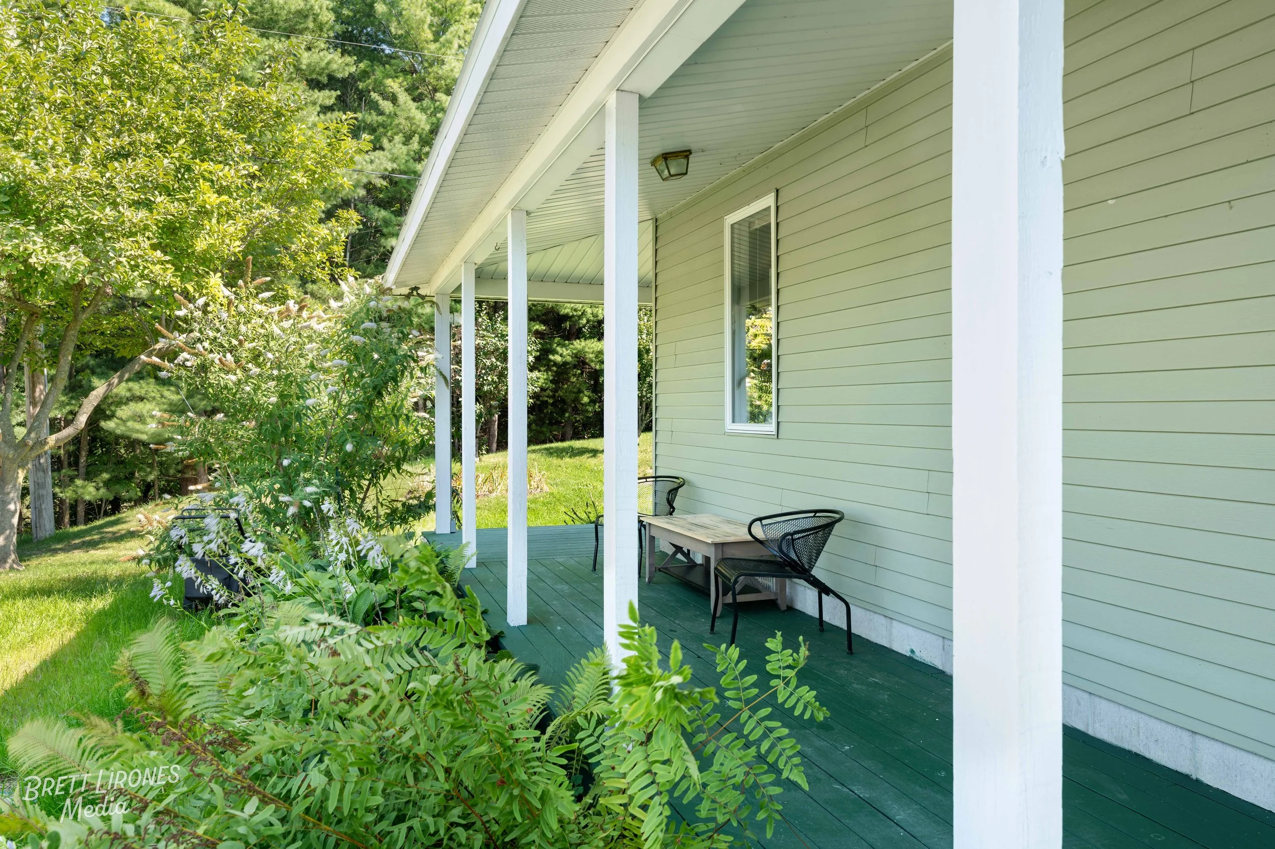 A porch with two black chairs and a small table, attached to a yellow house with white trim, surrounded by green trees and grass.