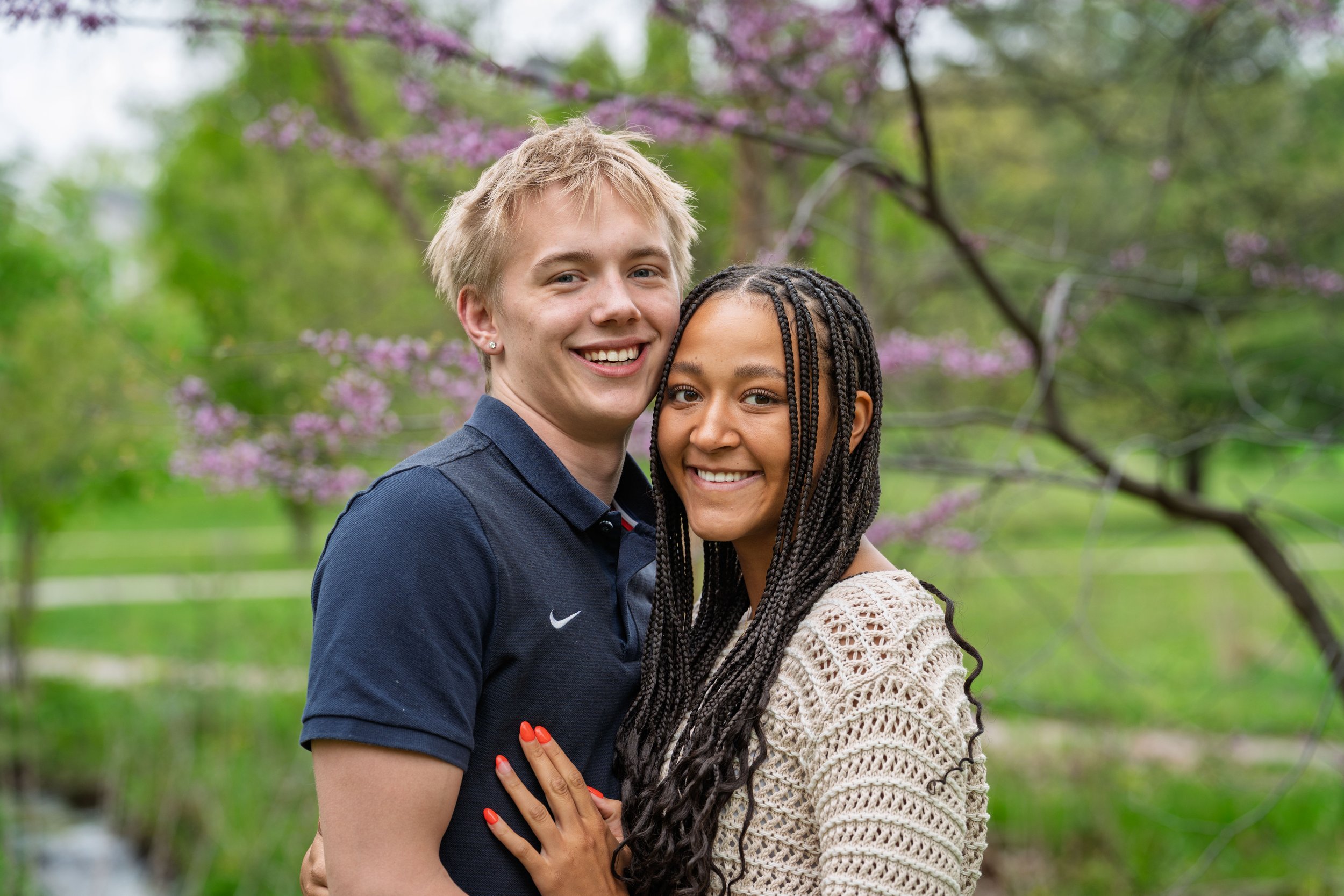 A smiling young man with blond hair and a young woman with braided hair stand close together outdoors with trees and pink blossoms in the background.