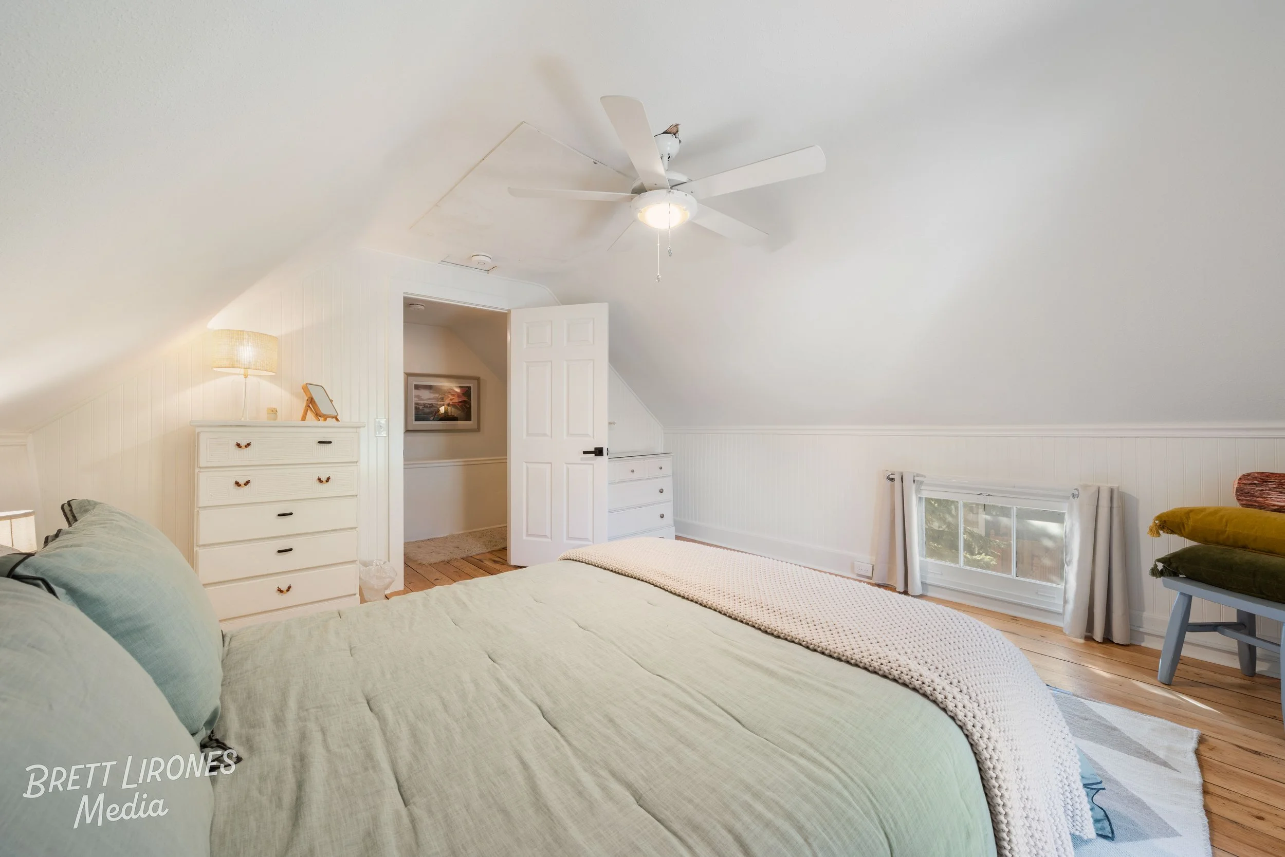 A cozy bedroom with a sloped ceiling, featuring a bed with light green bedding, a white dresser, and a small bench with stacked pillows near a window with curtains.