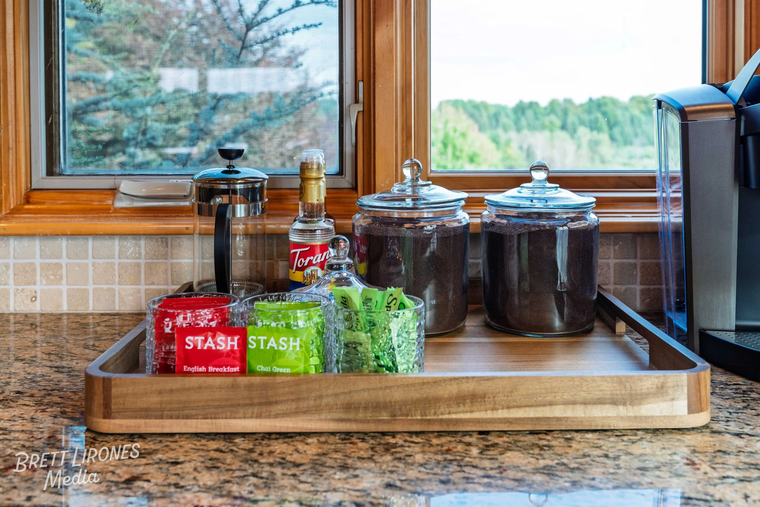 Kitchen countertop with glass jars of coffee, a bottle of Torani syrup, a Keurig coffee maker, and packets of tea in a wooden tray, with windows showing a natural outdoor view.