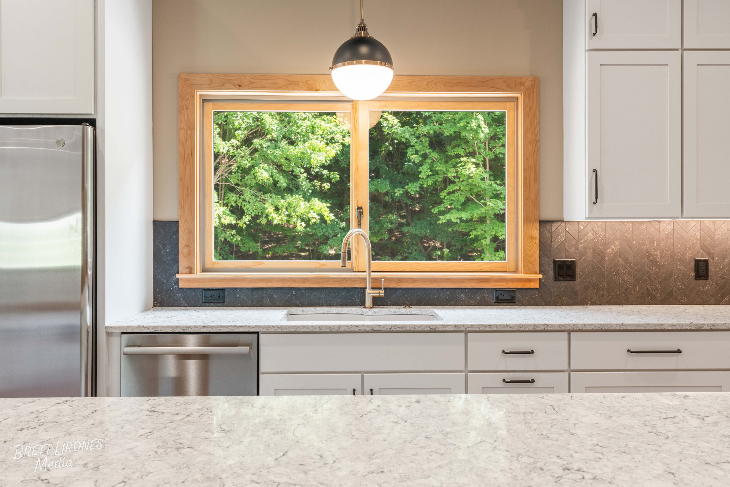 Modern kitchen with white cabinets, granite countertop, stainless steel appliances, and large window showing green trees outside.