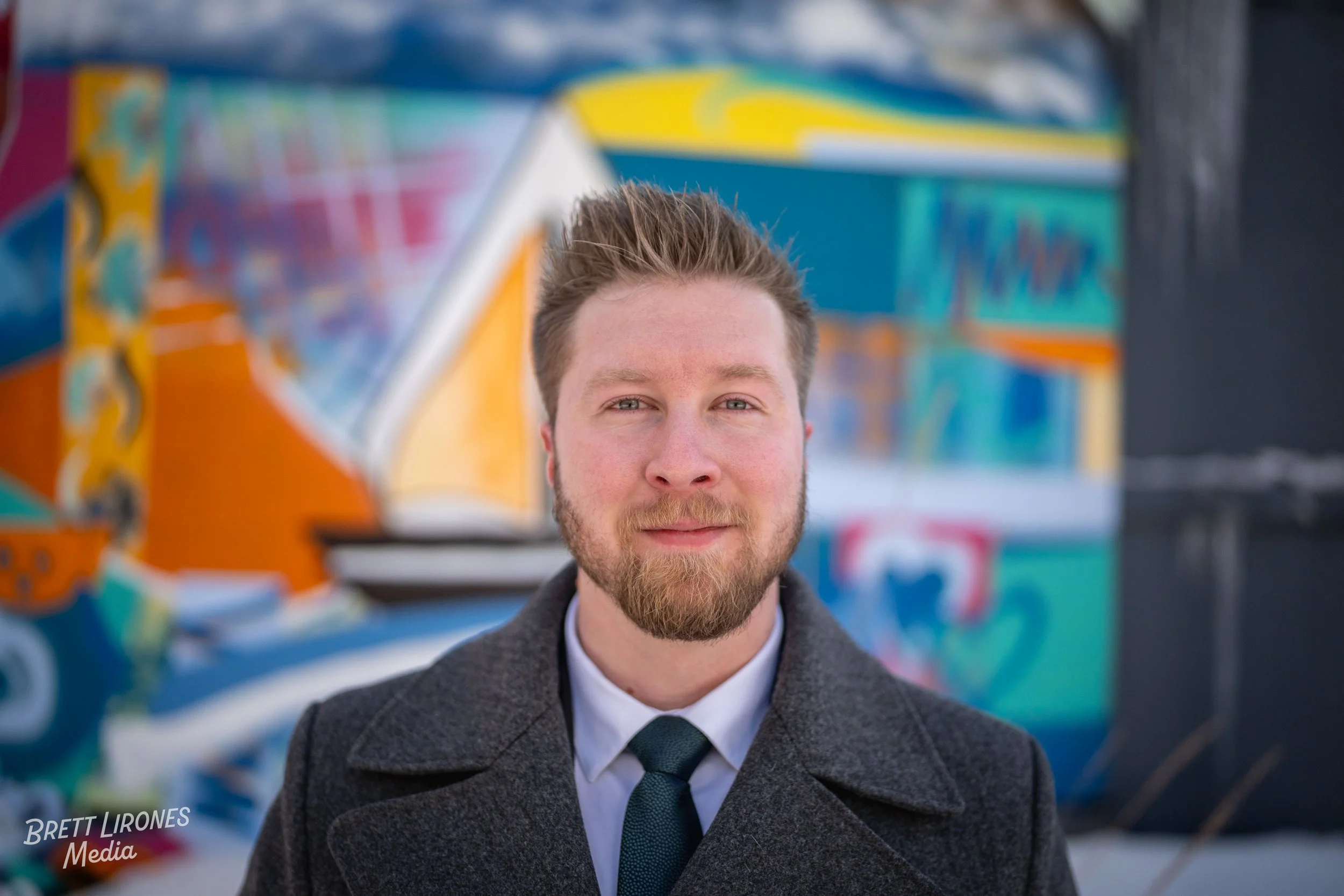 Portrait of a young man with light skin, light brown hair, and a beard, wearing a dark gray coat and a dark tie, standing in front of a colorful graffiti mural.