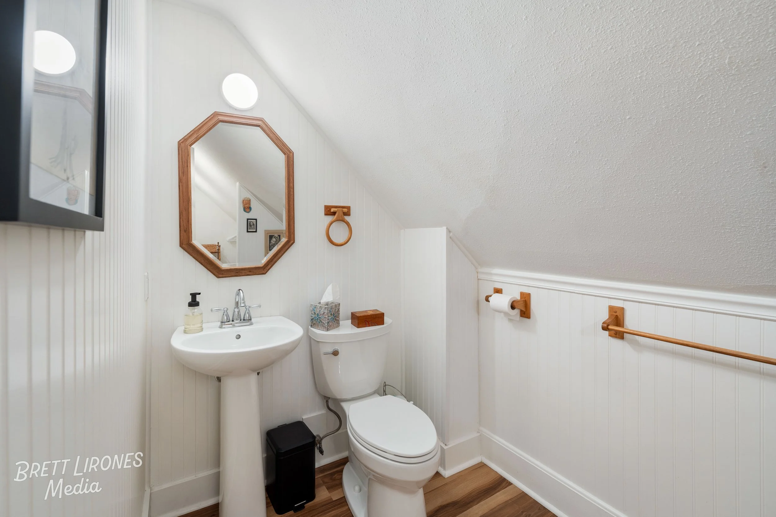 Small bathroom with a toilet, a pedestal sink, a mirror with a wooden frame, and wooden accessories including a towel ring, tissue box, and toilet paper holder. The walls are white beadboard paneling and the ceiling is sloped.