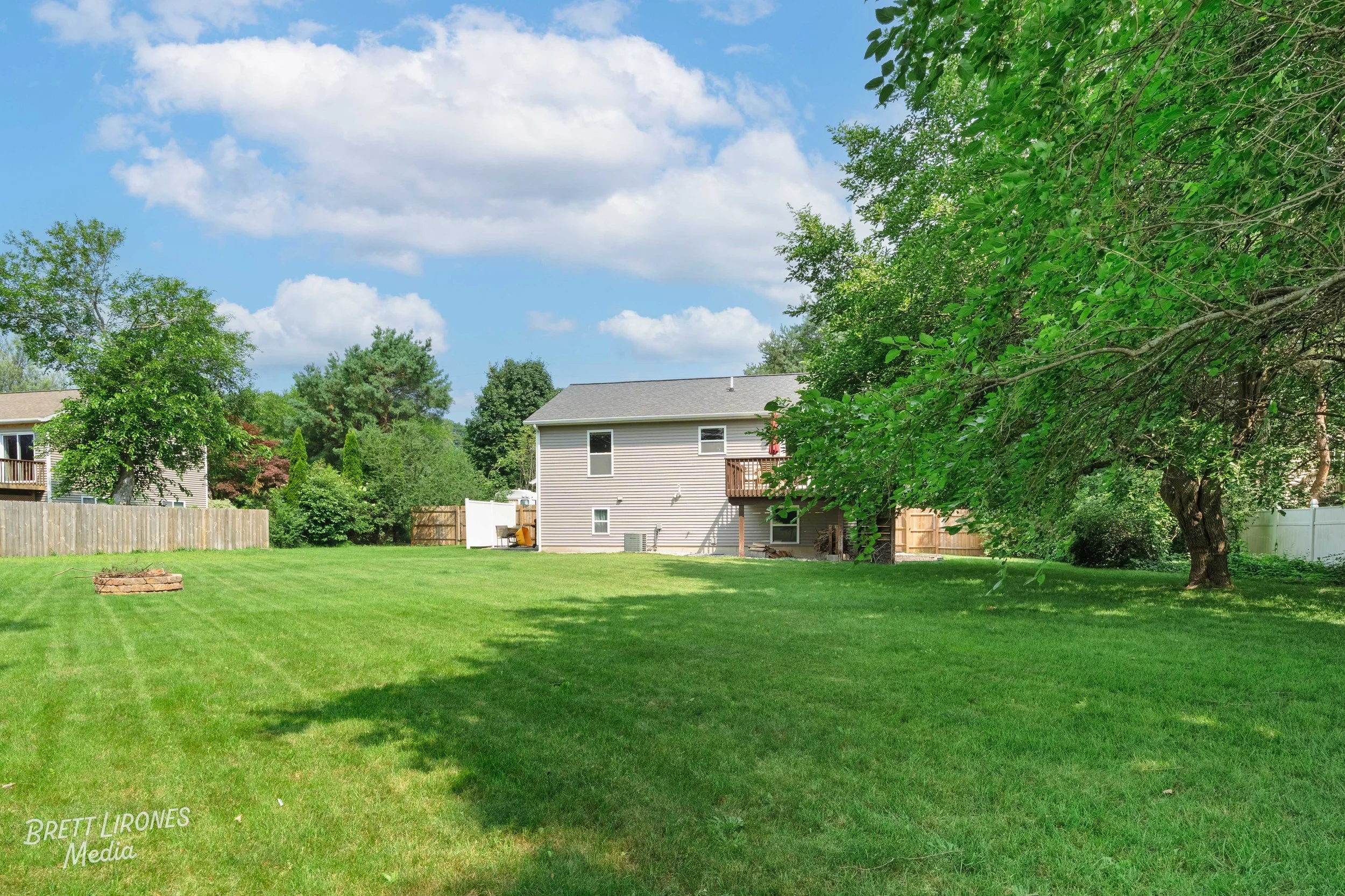 View of a backyard with a large green grassy lawn, mature trees on the right, a gray house with a deck in the background, and a blue sky with scattered clouds.