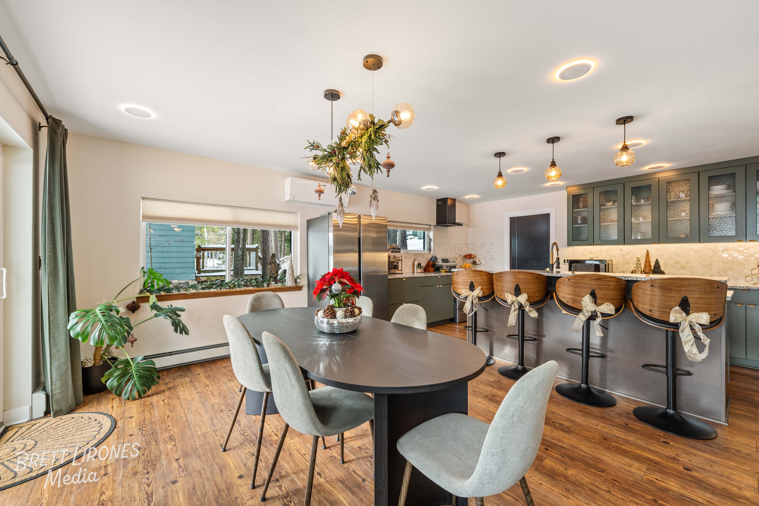 A modern kitchen and dining area with a dark wood oval table surrounded by six light gray upholstered chairs and a poinsettia centerpiece. Large windows with light-colored blinds, dark green cabinets with glass-fronted doors, a stainless steel refrig