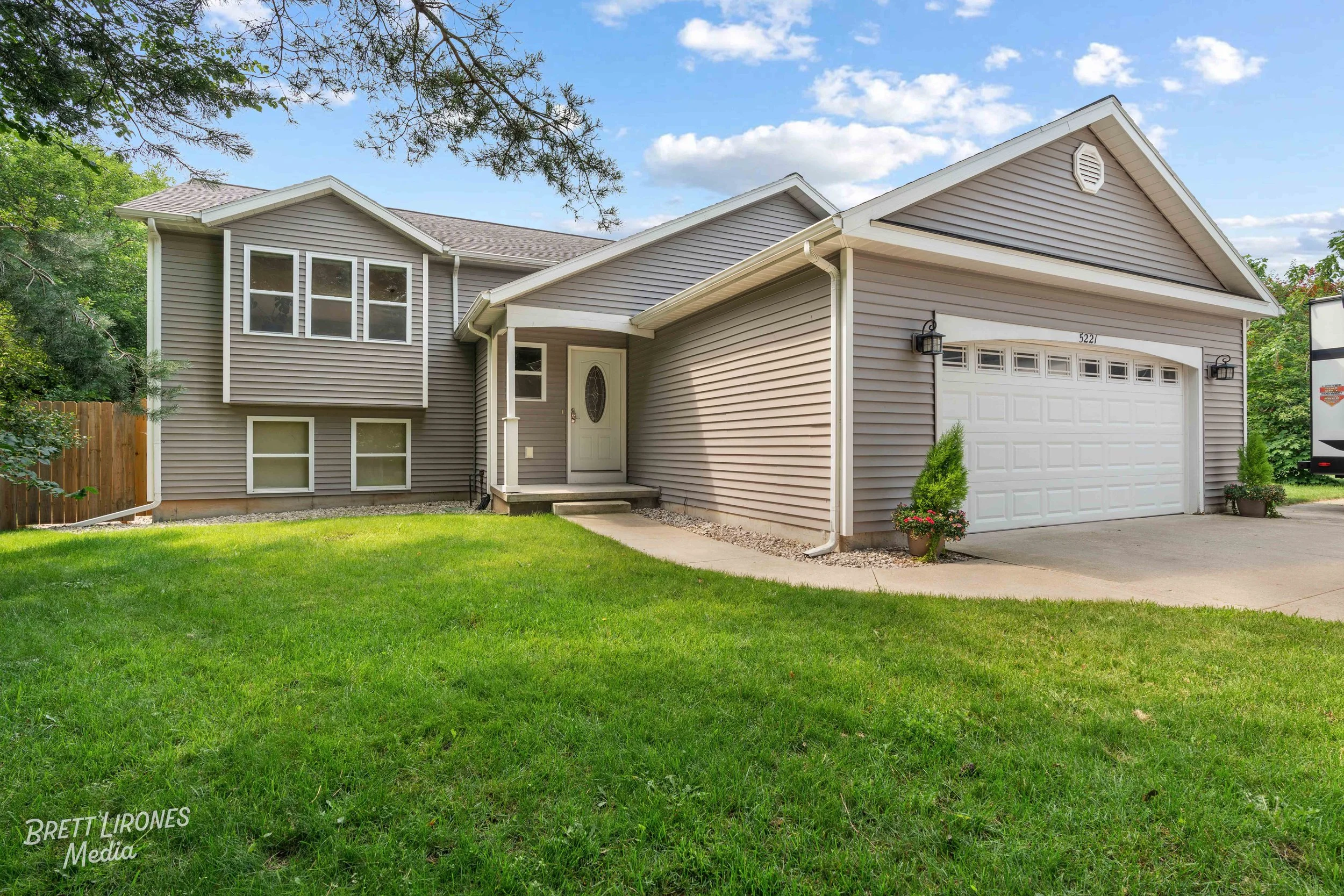 Front view of a beige house with a two-car garage and a well-maintained lawn, under a blue sky with some clouds.