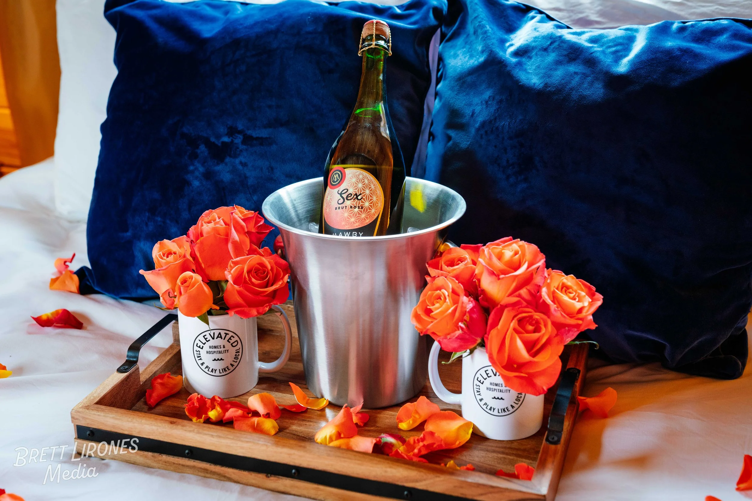 A bed with black and white pillows, a wooden tray holding two white mugs with orange roses, a metal ice bucket with a bottle labeled 'Sex' in the center, and scattered orange rose petals.