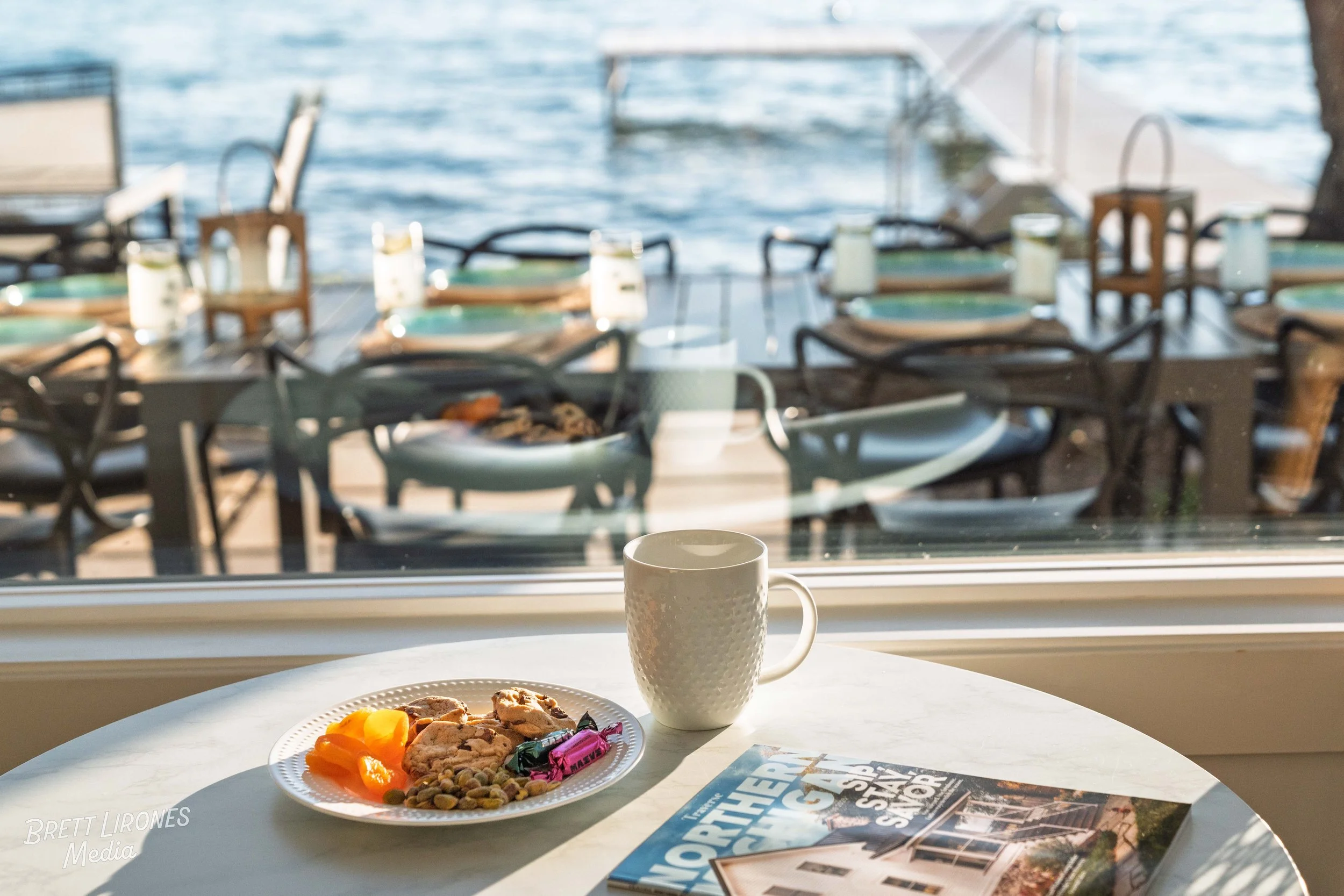 A breakfast table with cookies, dried apricots, pistachios, wrapped candy, a white mug, and a magazine near a window overlooking a dock with tables, chairs, and a body of water.