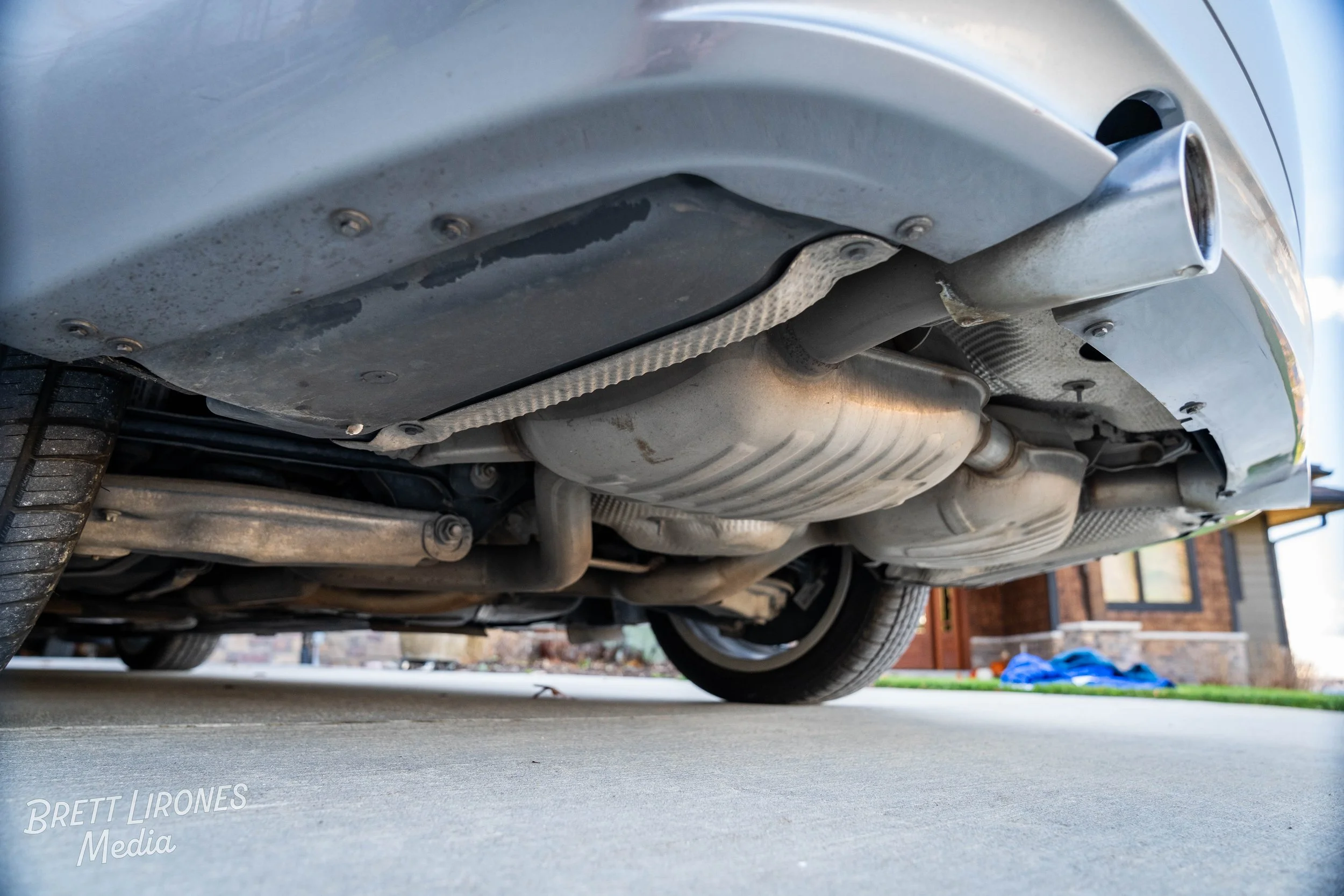 Underneath view of a silver car showing exhaust system and part of rear tire on concrete driveway with house in background.