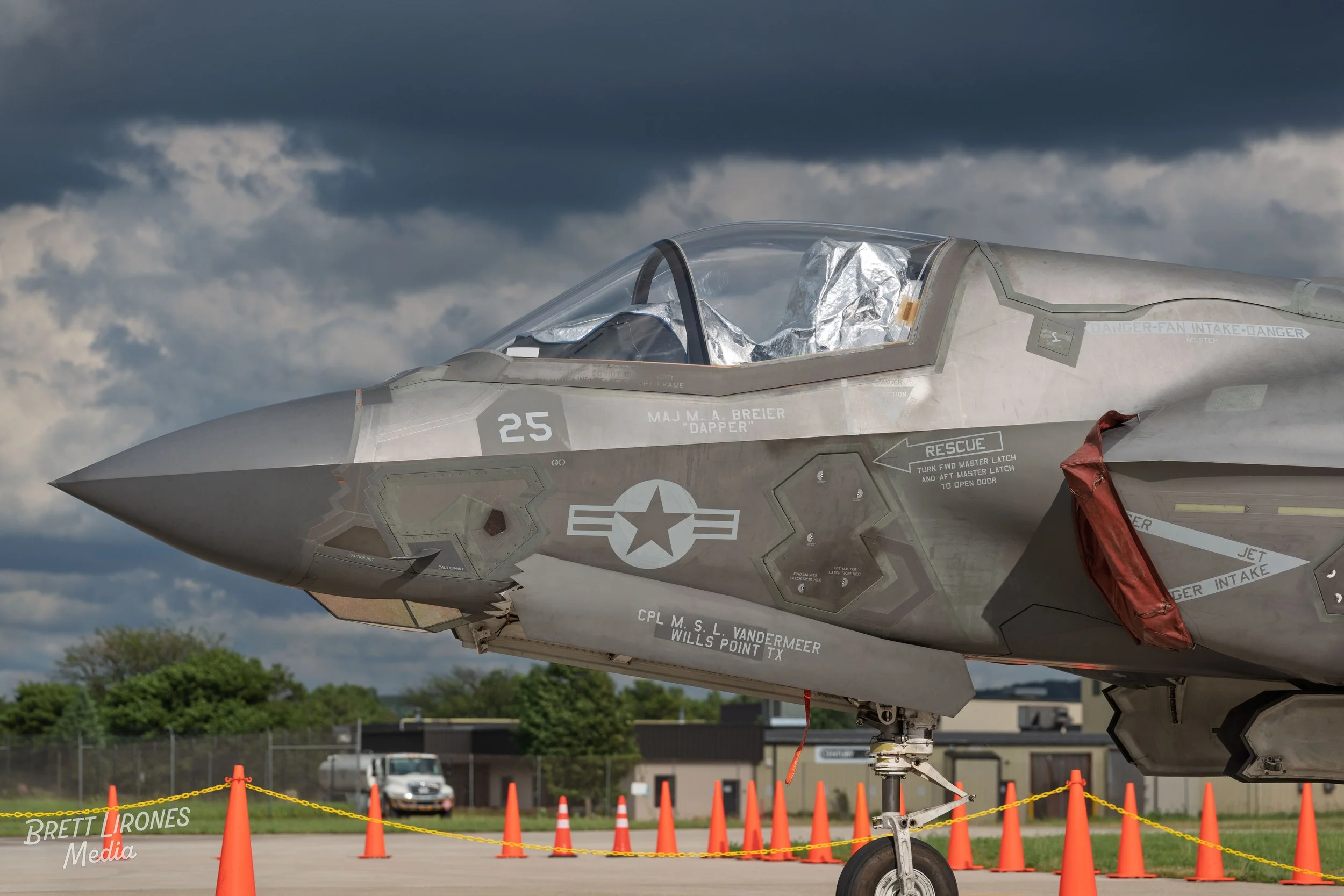 A fighter jet aircraft parked on the ground, marked with military insignia and safety instructions, surrounded by orange traffic cones and chains.