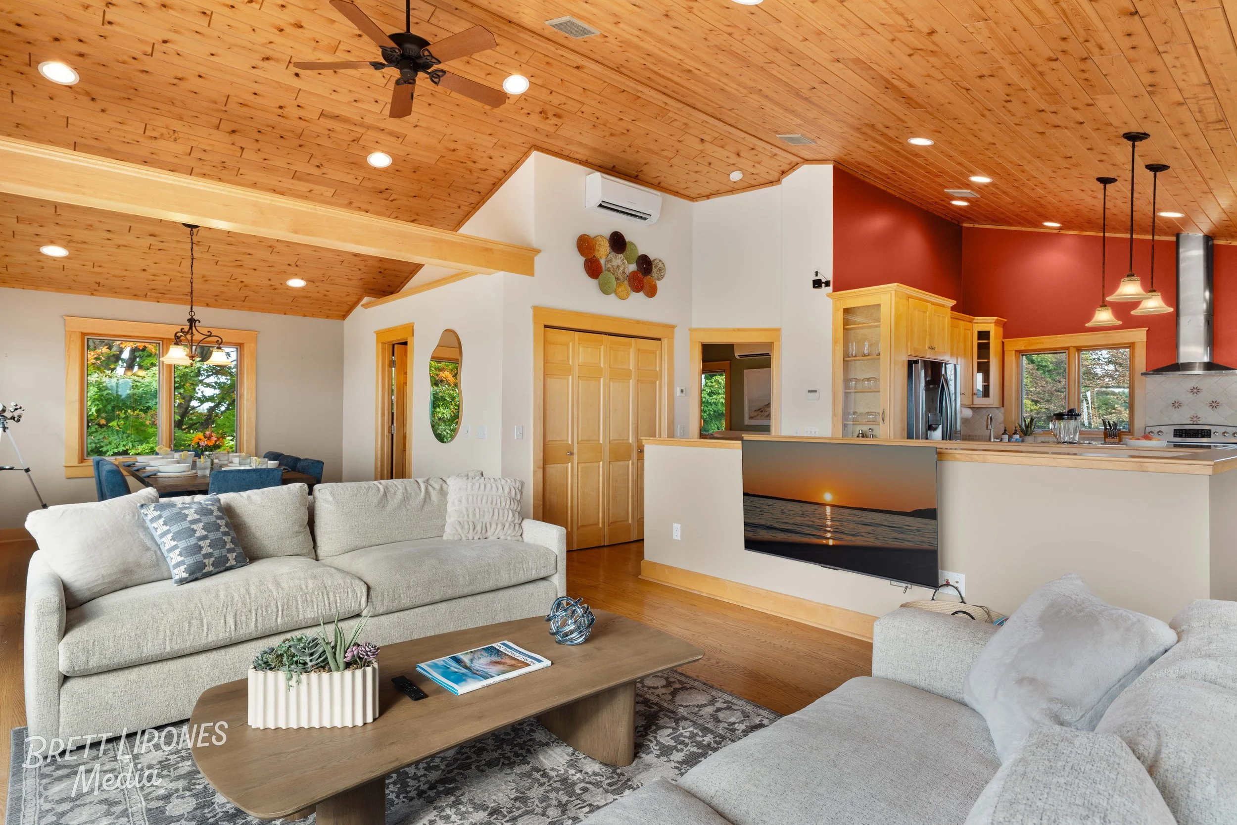 Living room with beige sofas, wooden coffee table, and a wall-mounted TV showing a sunset over water. The room has a wood-paneled ceiling with ceiling fan and recessed lighting. In the background is a dining area and open kitchen with wooden cabinets