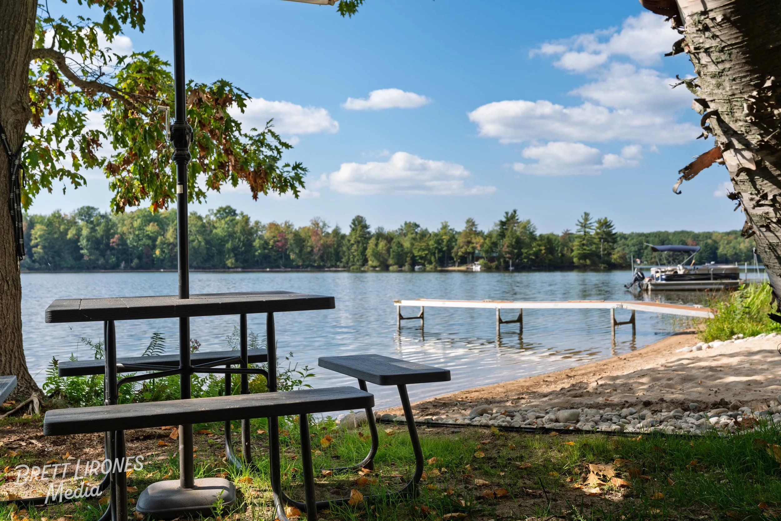 A lakeside scene with a picnic table and bench in the foreground, a dirt and pebble beach, a dock in the water, and boats docked on the opposite shore, with trees and a blue sky with clouds in the background.