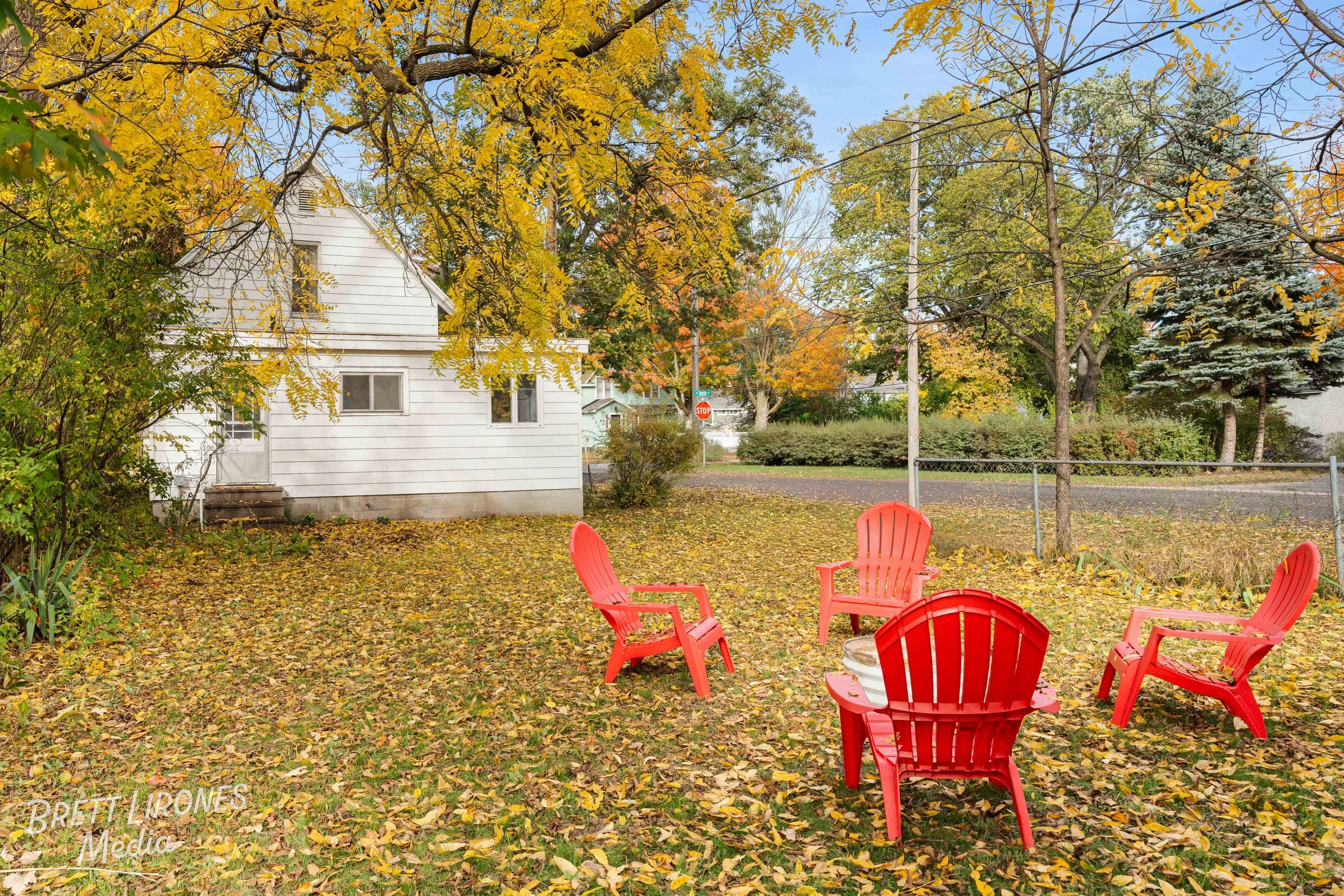A backyard scene during autumn with four red Adirondack chairs arranged in a circle on a leaf-covered lawn. There are trees with fall foliage surrounding the yard, and a white house is visible in the background. A stop sign and utility pole are at th