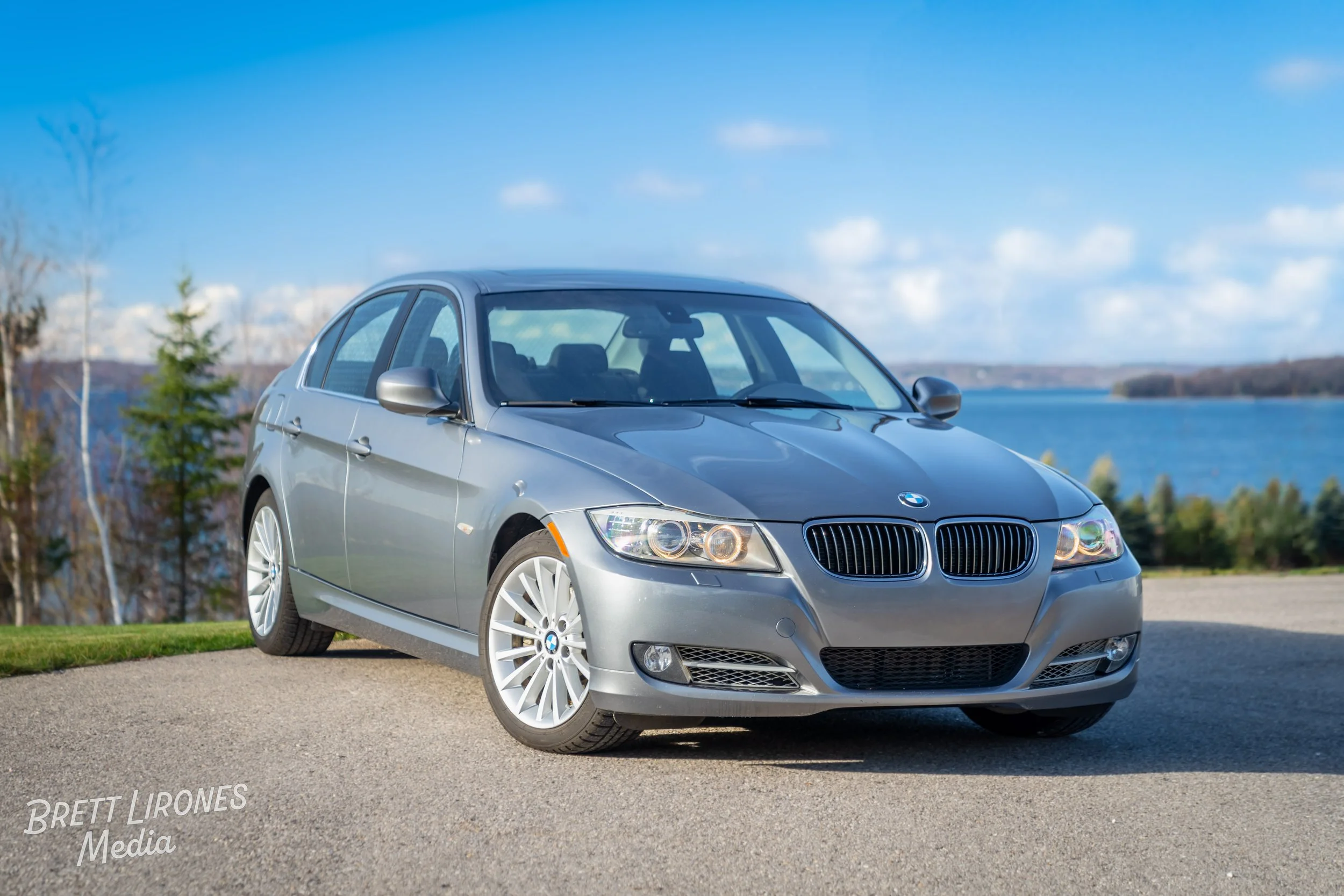 A silver BMW sedan parked on a paved road near a lake with trees and a blue sky in the background.