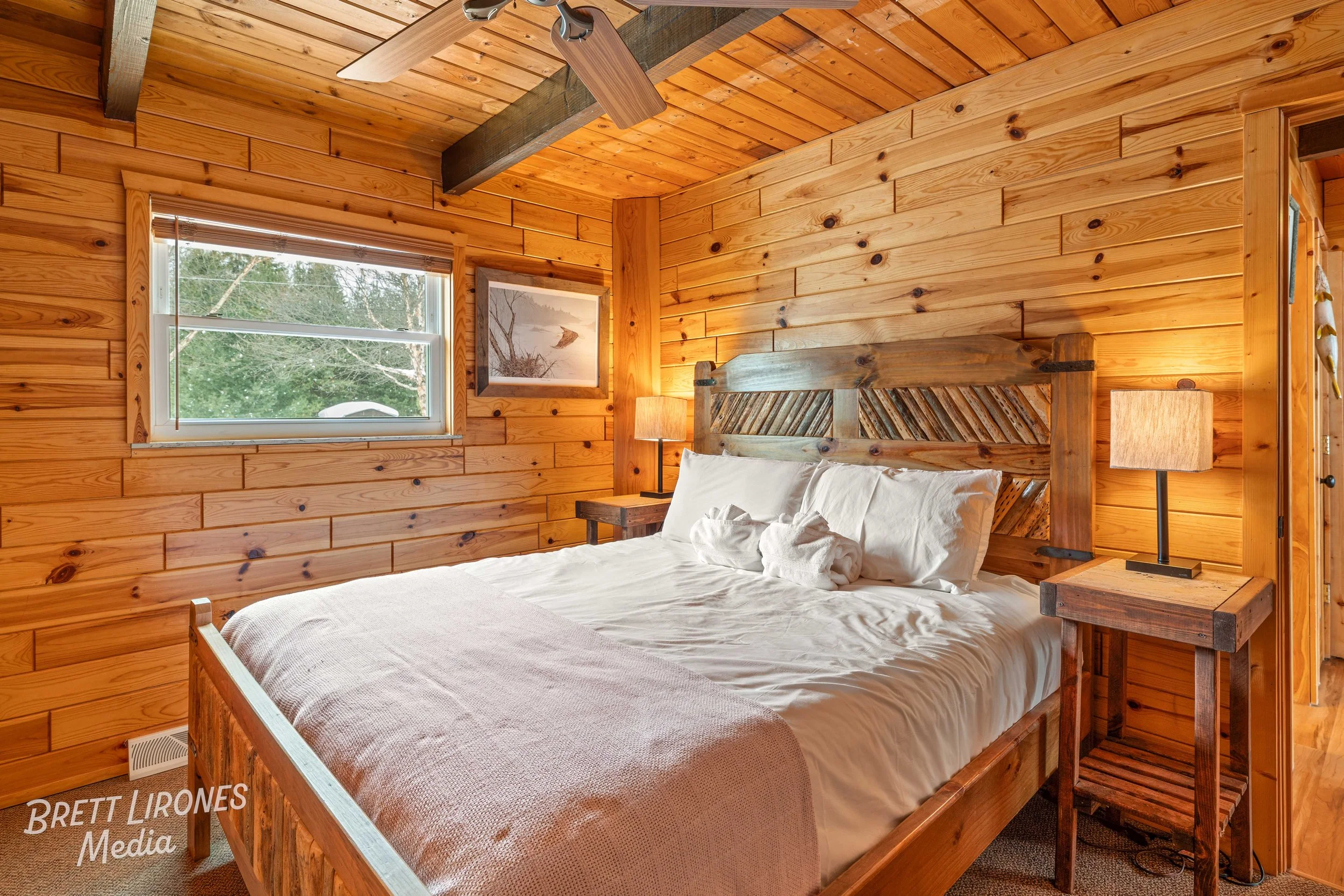 Rustic bedroom with wooden walls, twin nightstands with lamps, a large bed with white bedding, and a window showing greenery outside.