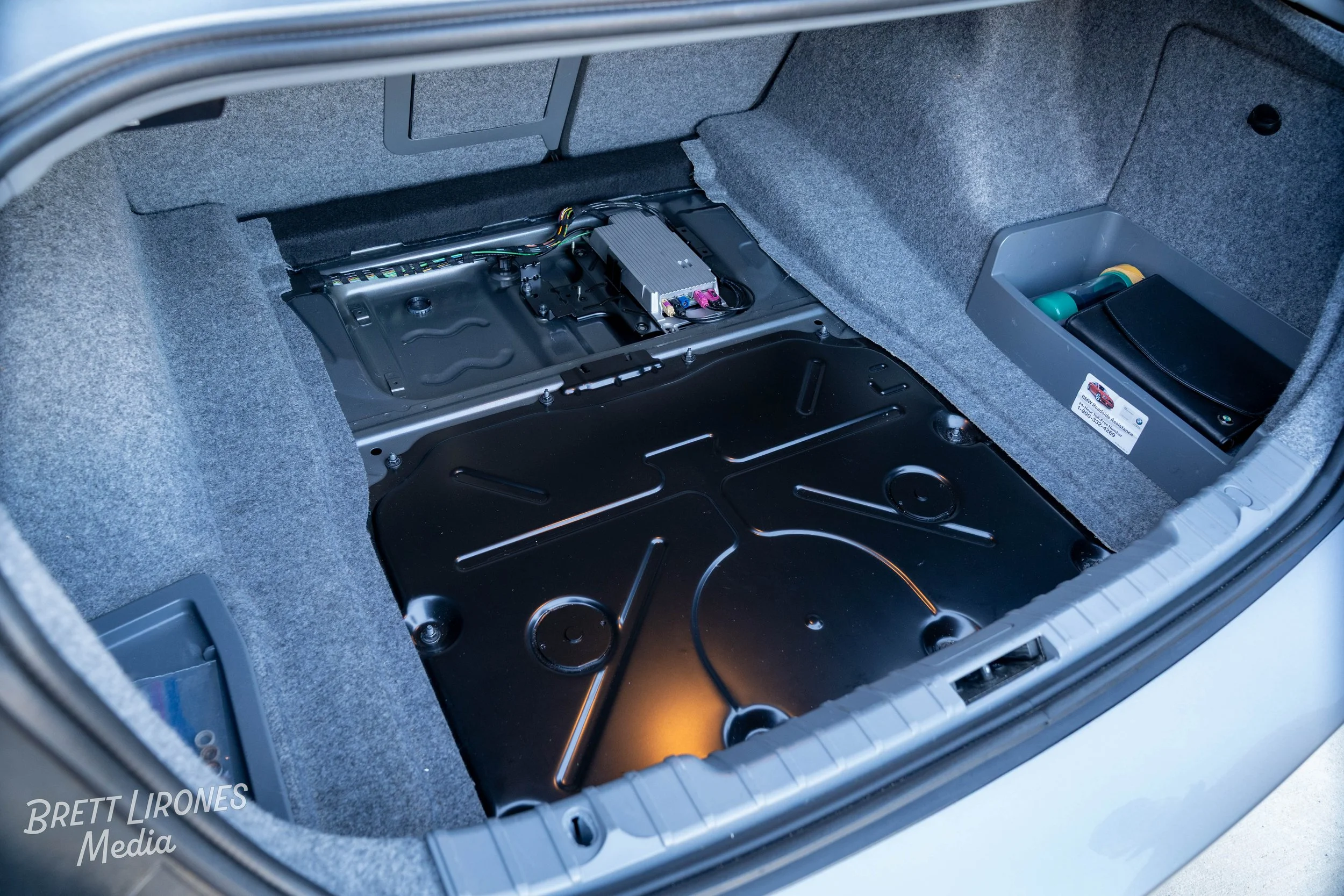 The trunk of a car with the interior panel removed, showing electronic components and wiring, with storage compartment on the side.