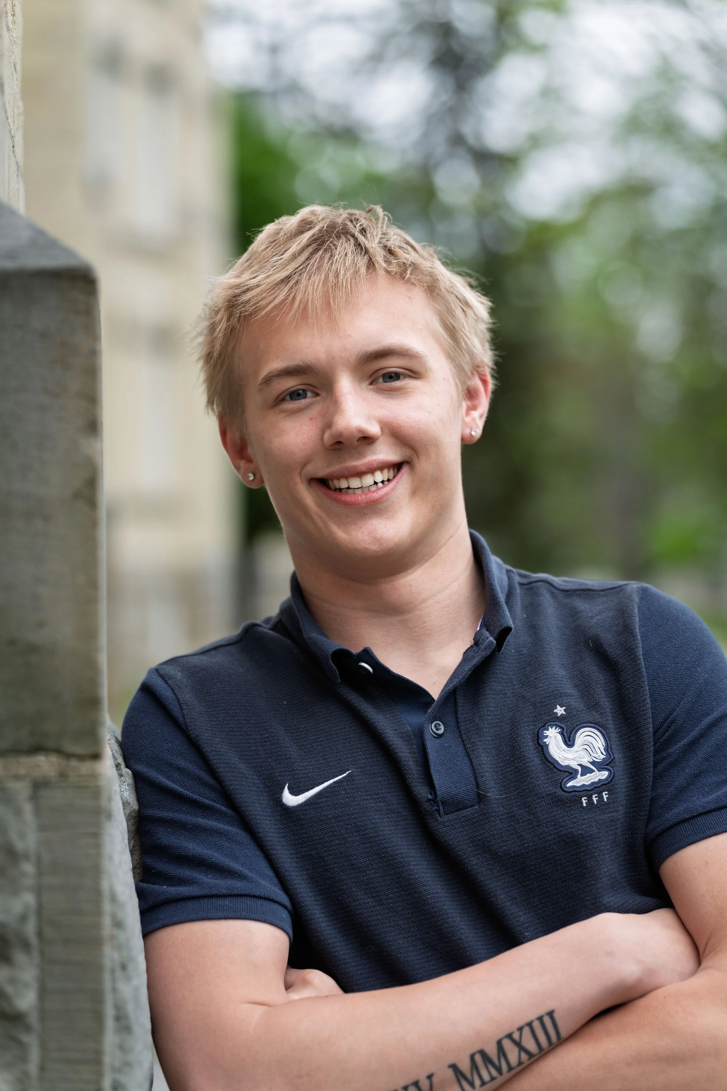 Young man smiling outdoors, wearing a navy blue polo shirt with a French football emblem and the Nike logo, with crossed arms, standing near a stone wall, blurred trees in the background.