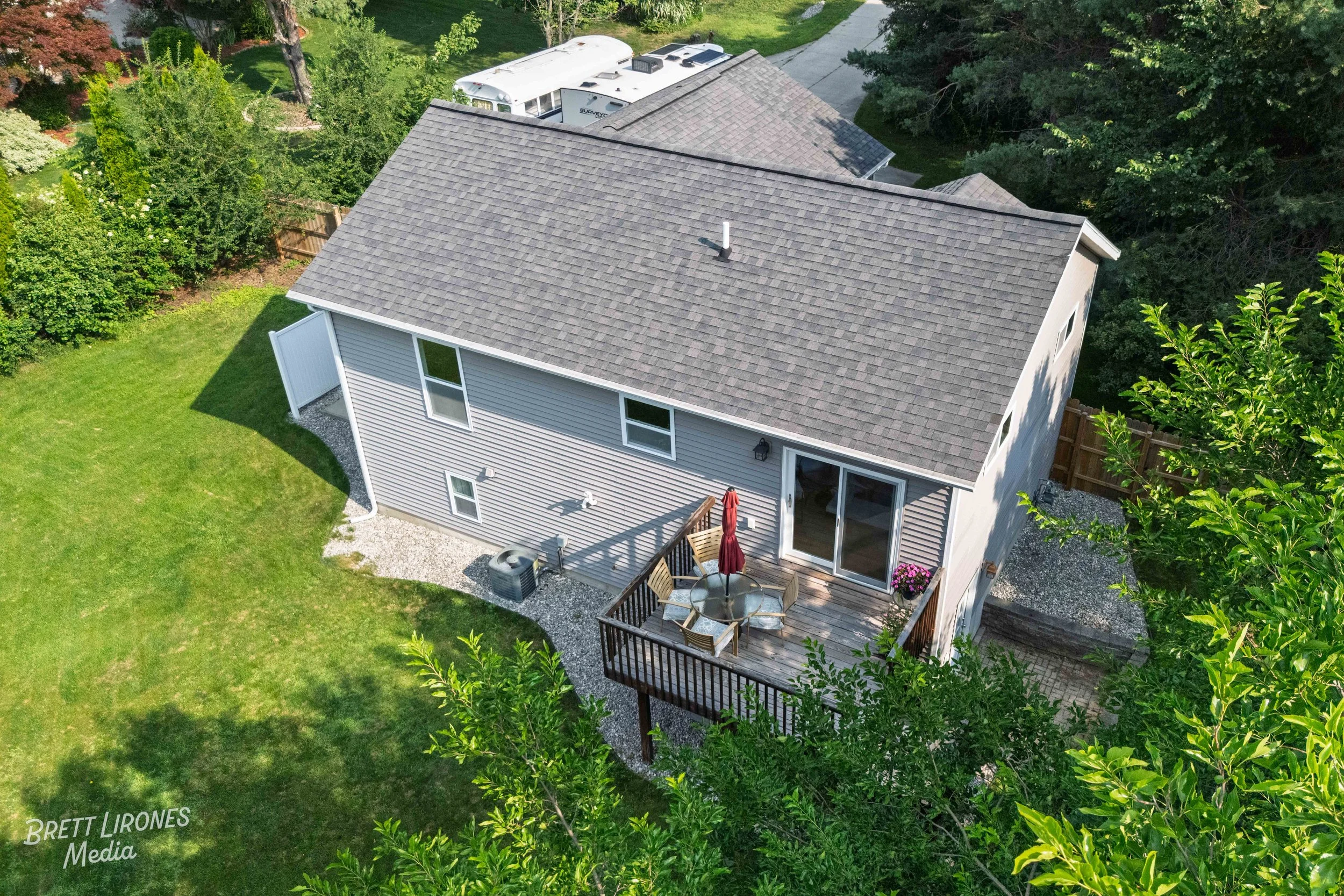 Aerial view of a backyard with a wooden deck, patio furniture with a red umbrella, sliding glass door, and lush green lawn surrounded by trees.