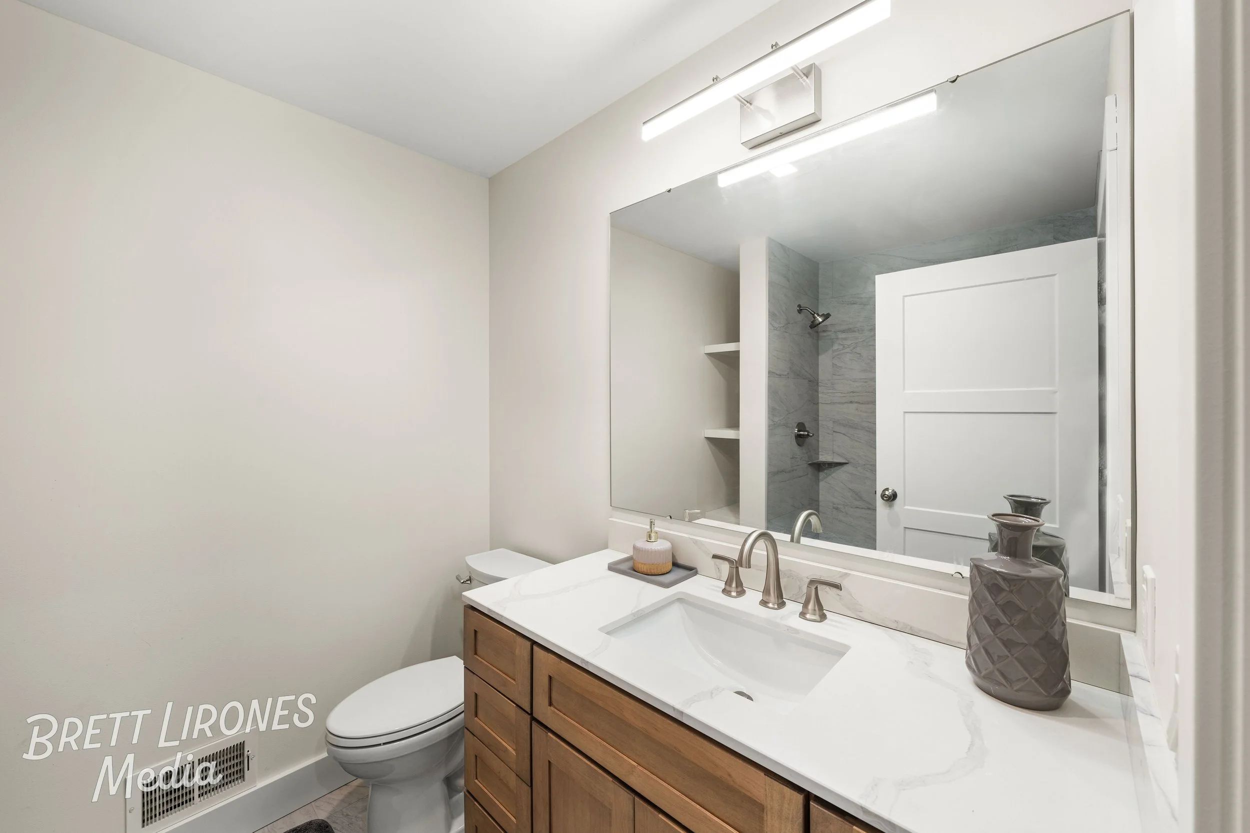 Bathroom with a large mirror, white marble countertop, wooden vanity, shower with gray marble walls, and decorative vases.