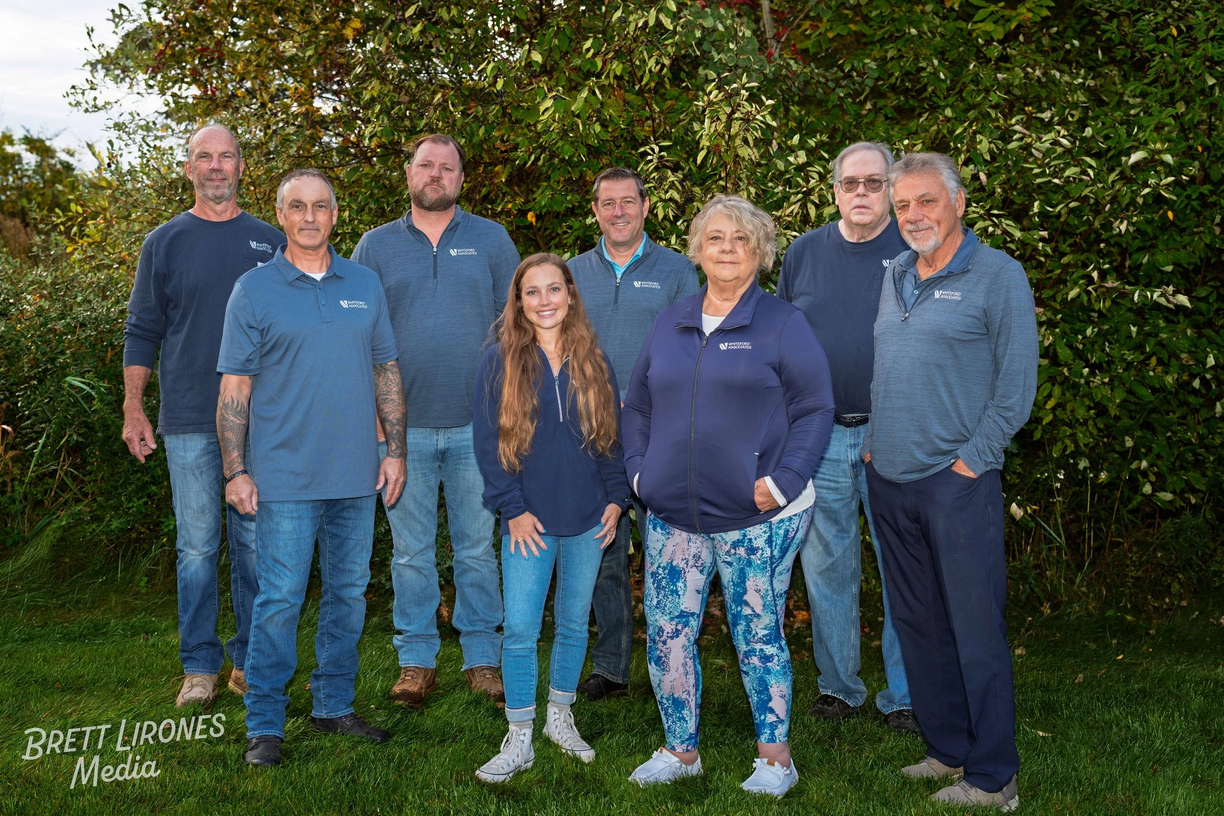 Group of nine people standing outdoors in front of green shrubbery, wearing blue and navy clothing, some with logo patches. The group includes men and women of various ages, all smiling or looking at the camera.