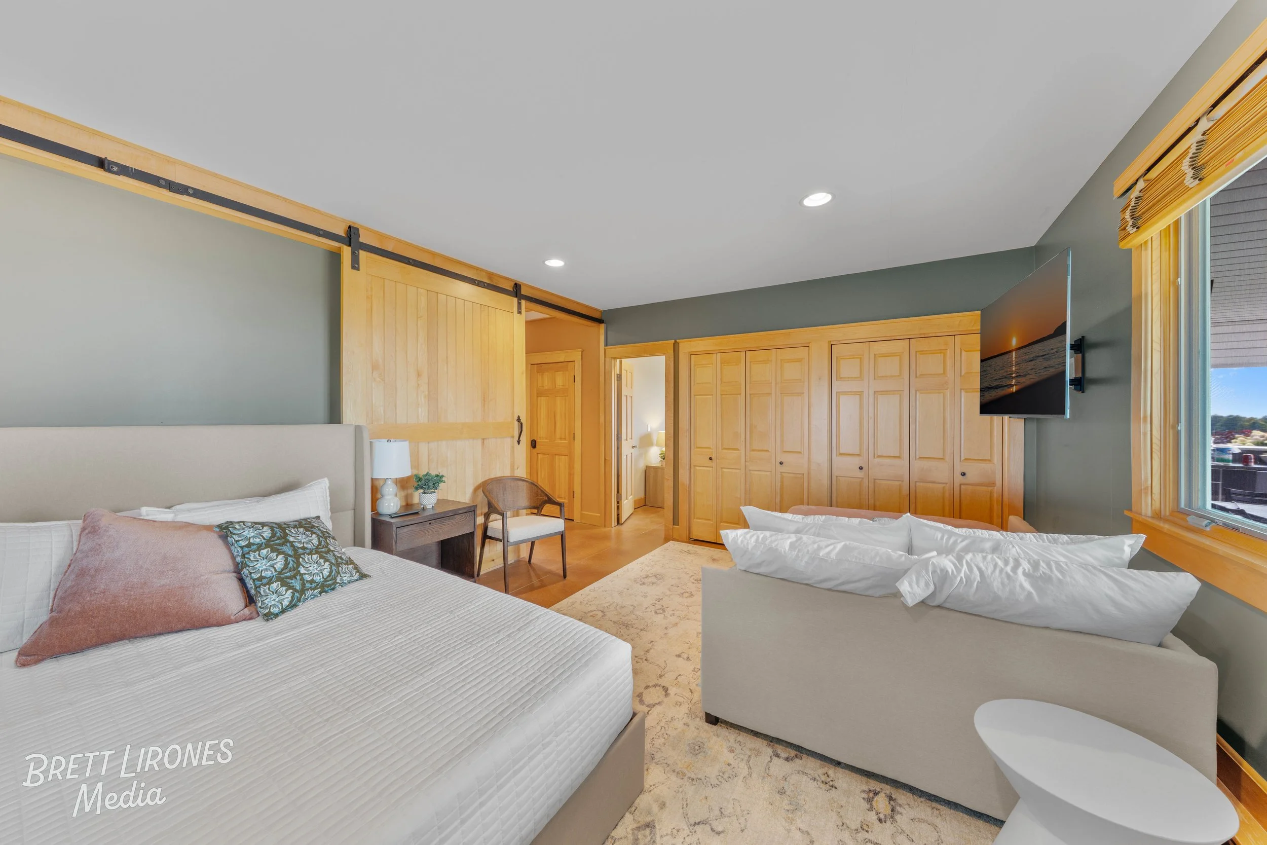 Interior of a bedroom with a large bed, a small side table with a lamp, a chair, a sofa, a wall-mounted TV, and multiple wooden closet doors, with a window showing an outdoor scene.