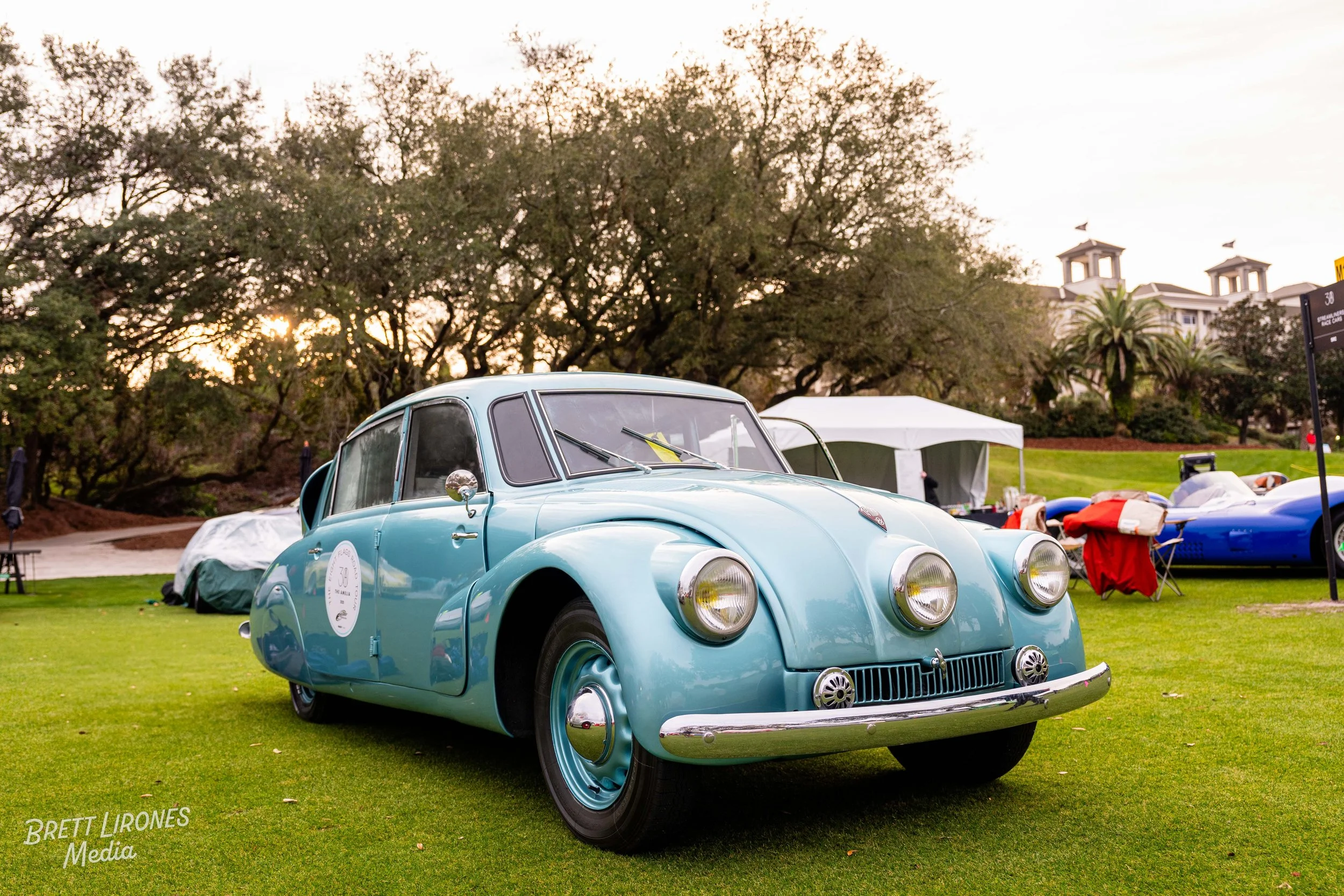 A vintage light blue car displayed on a grassy field during an outdoor car show, with other classic cars and tents visible in the background.