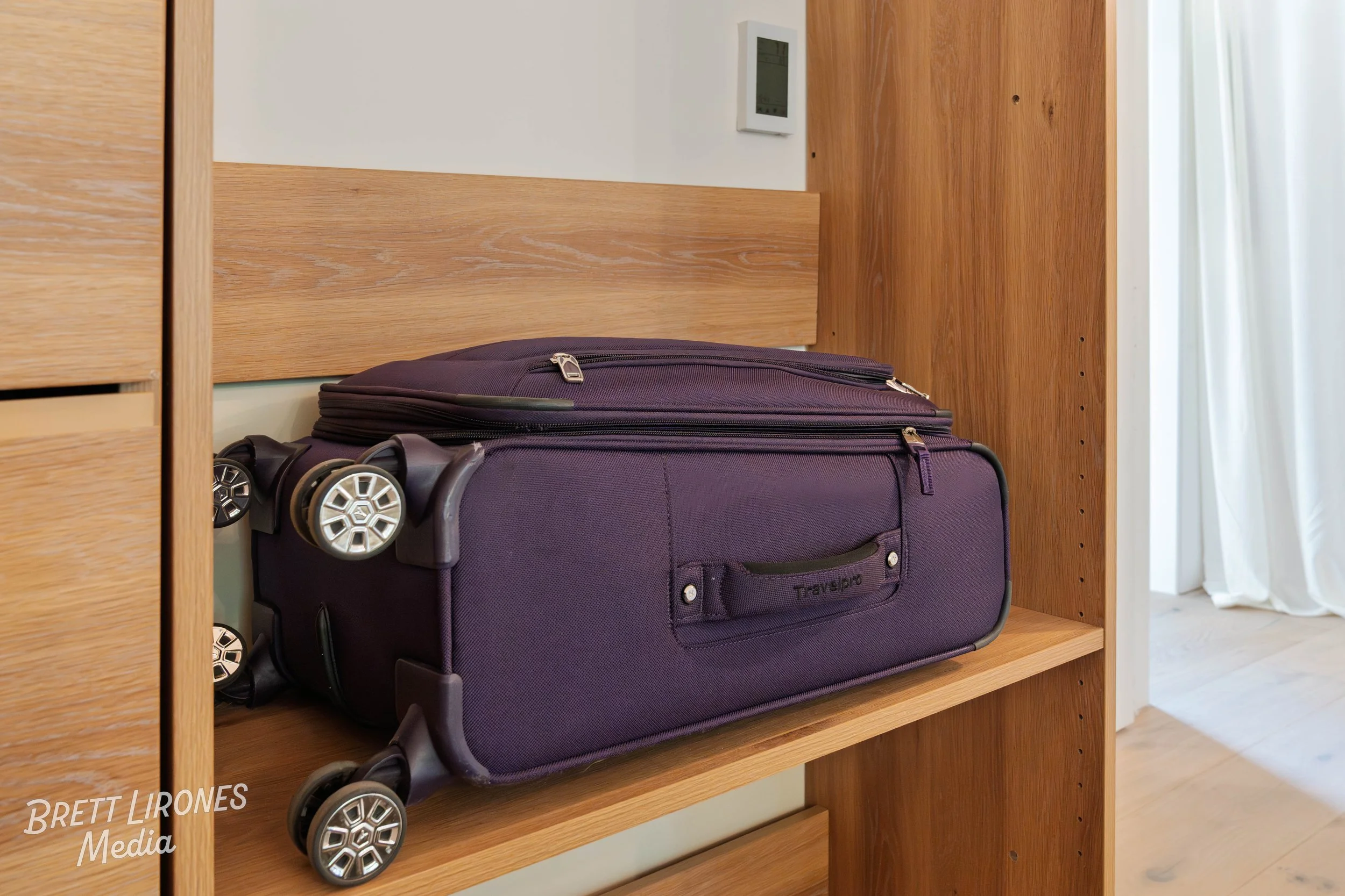A purple rolling suitcase lying on its side on a wooden shelf in a room with white walls and curtains.