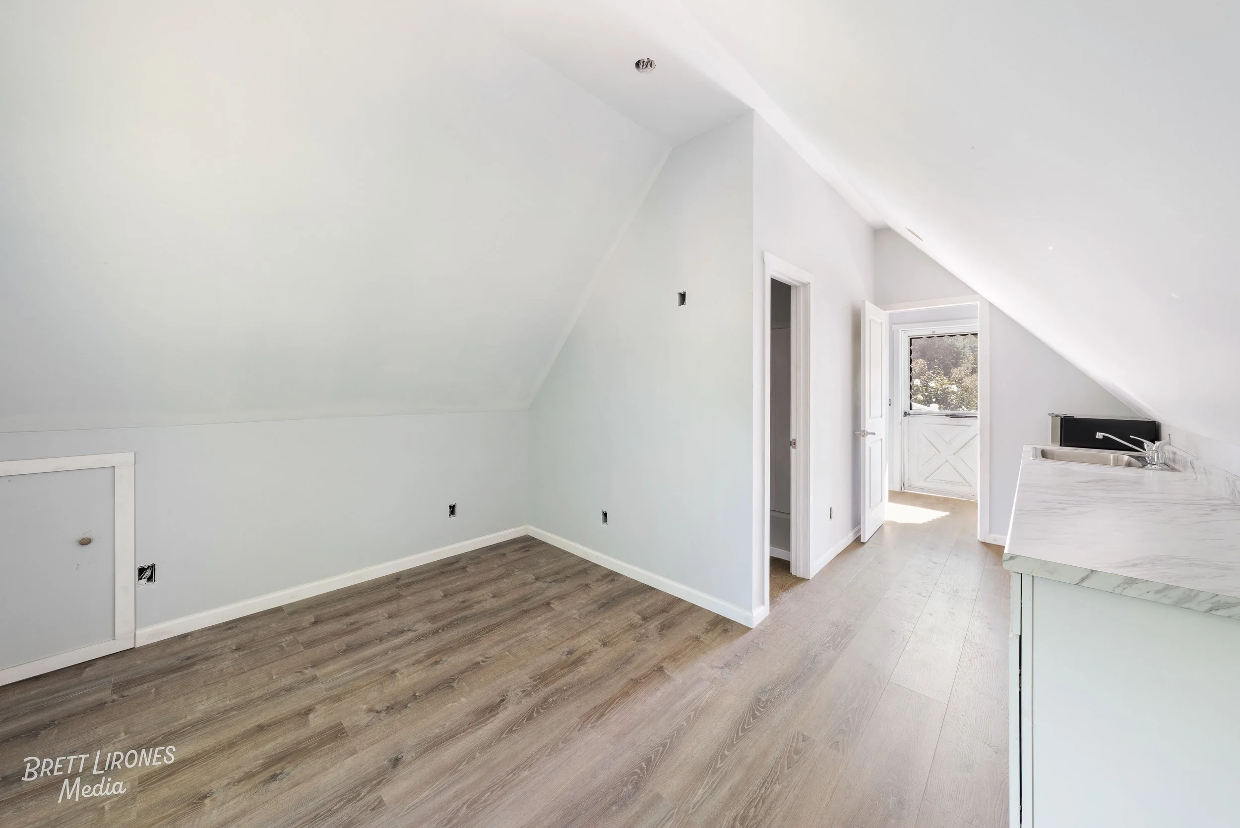 Empty room with sloped ceiling, light-colored walls, and wooden floor. A small door is visible on the left side. There is a doorway leading to outside with a view of trees, and part of a kitchen with a marble countertop is on the right.