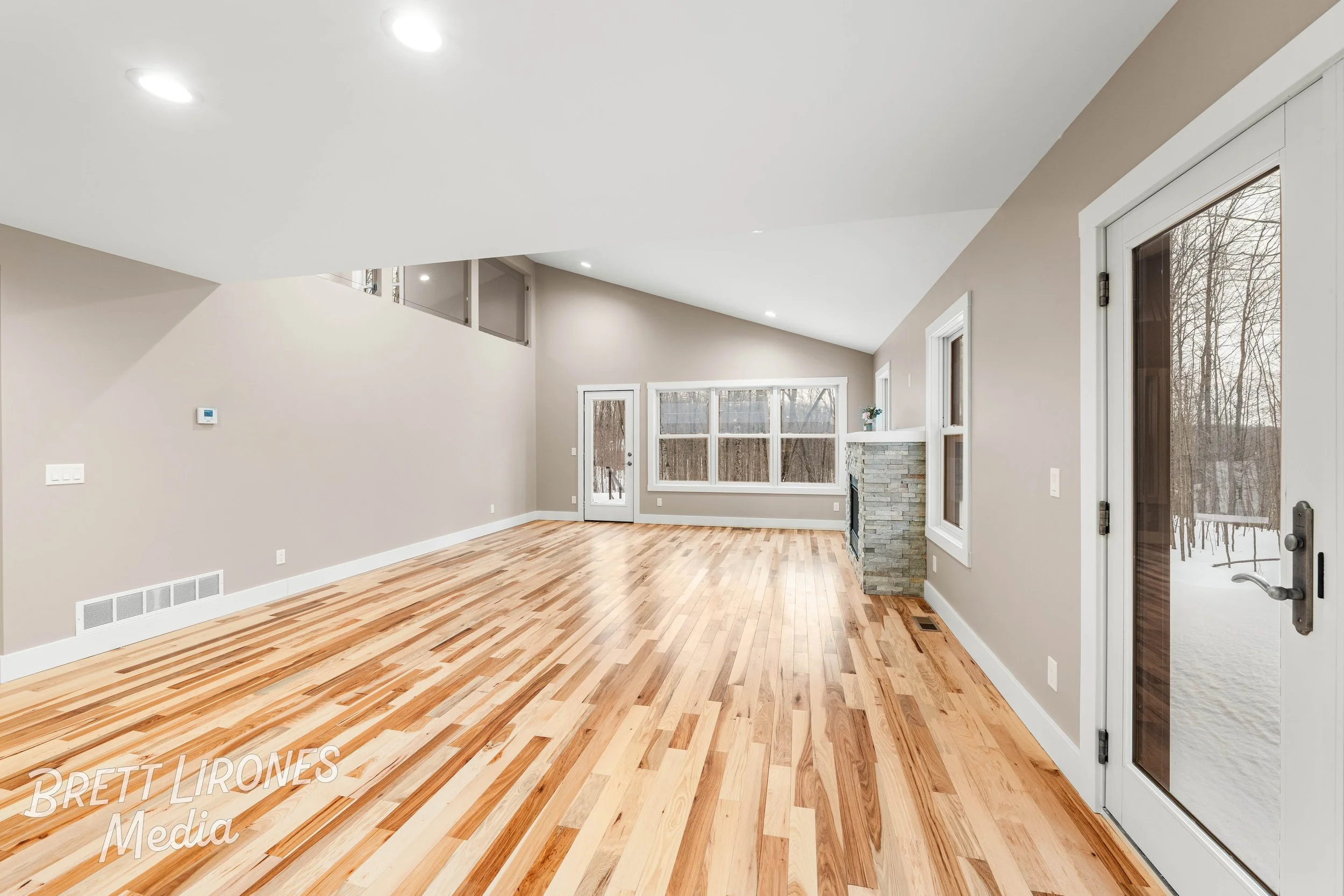 Empty living room with hardwood floors, beige walls, large windows, and a stone fireplace with a raised hearth, outside snow visible through the glass door.