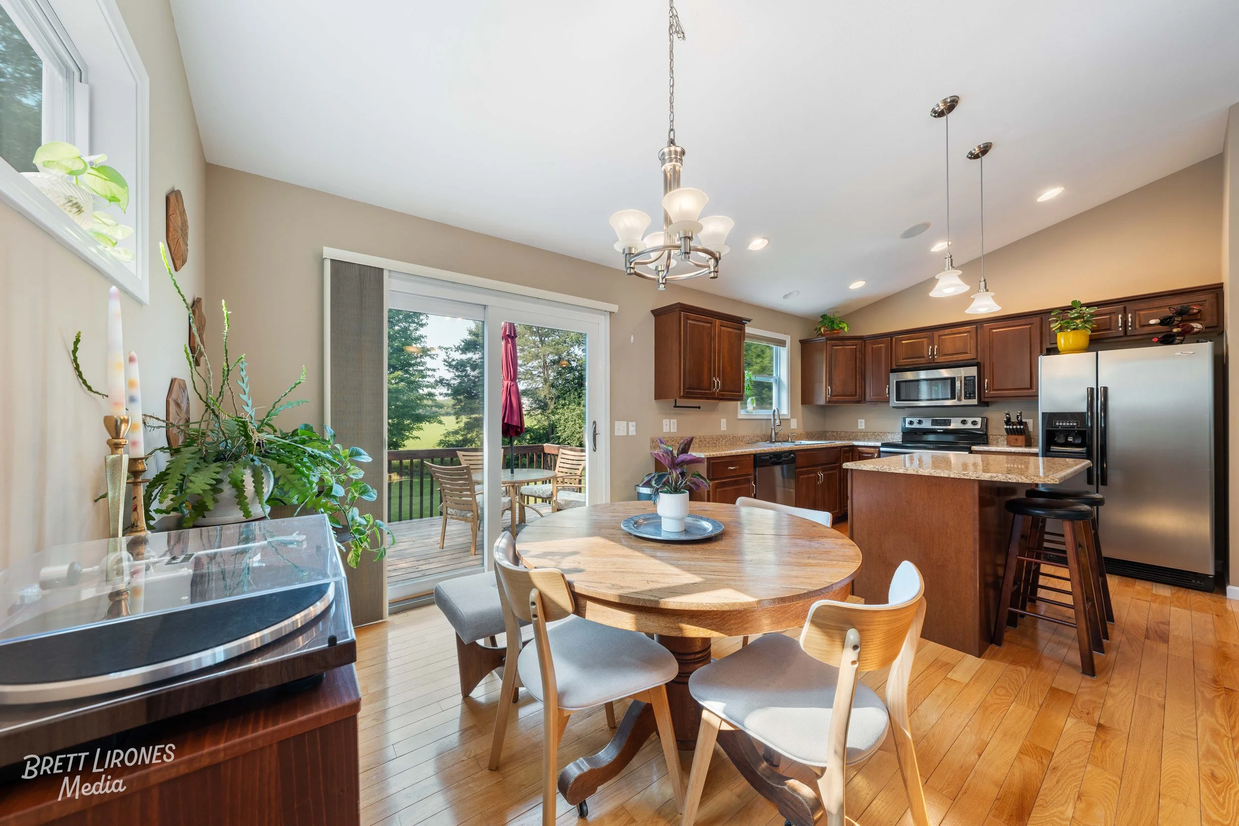 Kitchen and dining area with wooden floors, a round wooden table with four chairs, and a sliding glass door leading to an outdoor deck with patio furniture and greenery.
