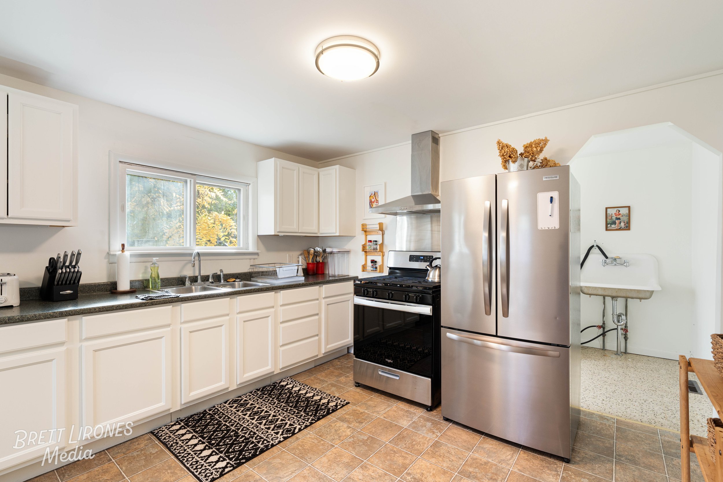 Kitchen with white cabinets, black countertop, stainless steel refrigerator, black stove, window above sink, dish drying rack, knife block, paper towel holder, small red containers, and sink area with soap dispenser.