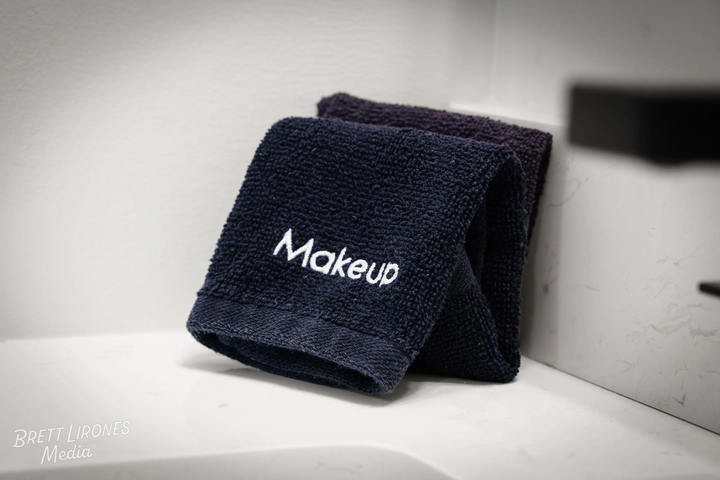A stack of black towels with white embroidered text that reads "Makeup" on the front towel, placed on a white shelf against a light-colored wall.