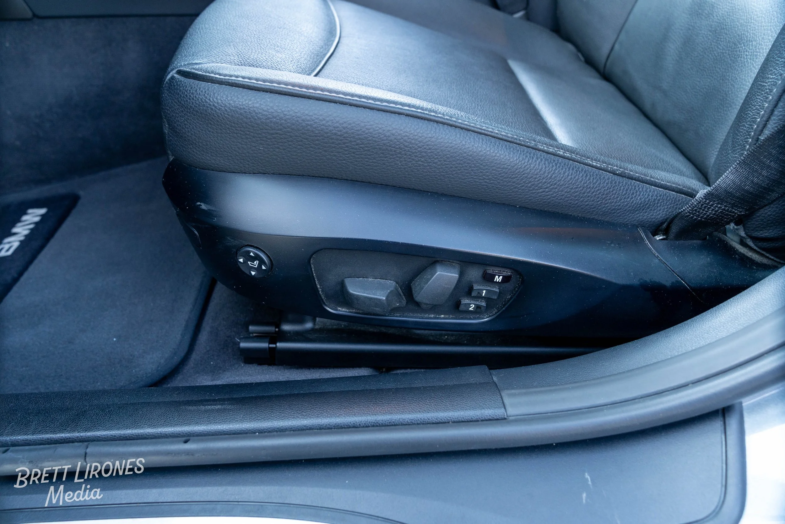 Close-up of a black leather car seat with electronic adjustment controls on the side, set on a dark gray car floor with a custom floor mat.