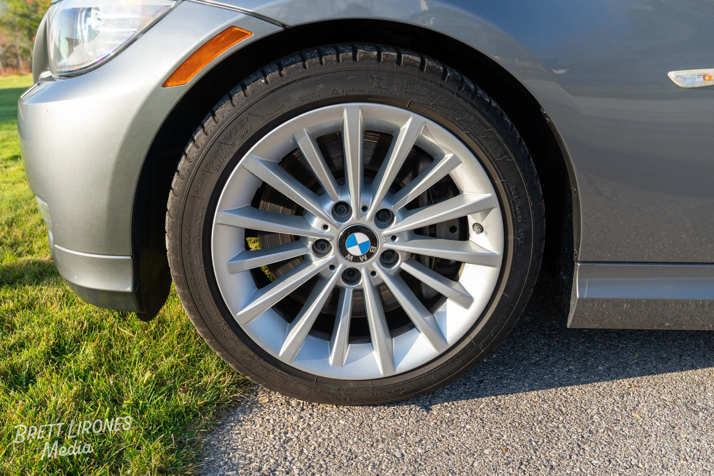 Close-up of a silver BMW car wheel and tire on a grey pavement with grass at the edge.