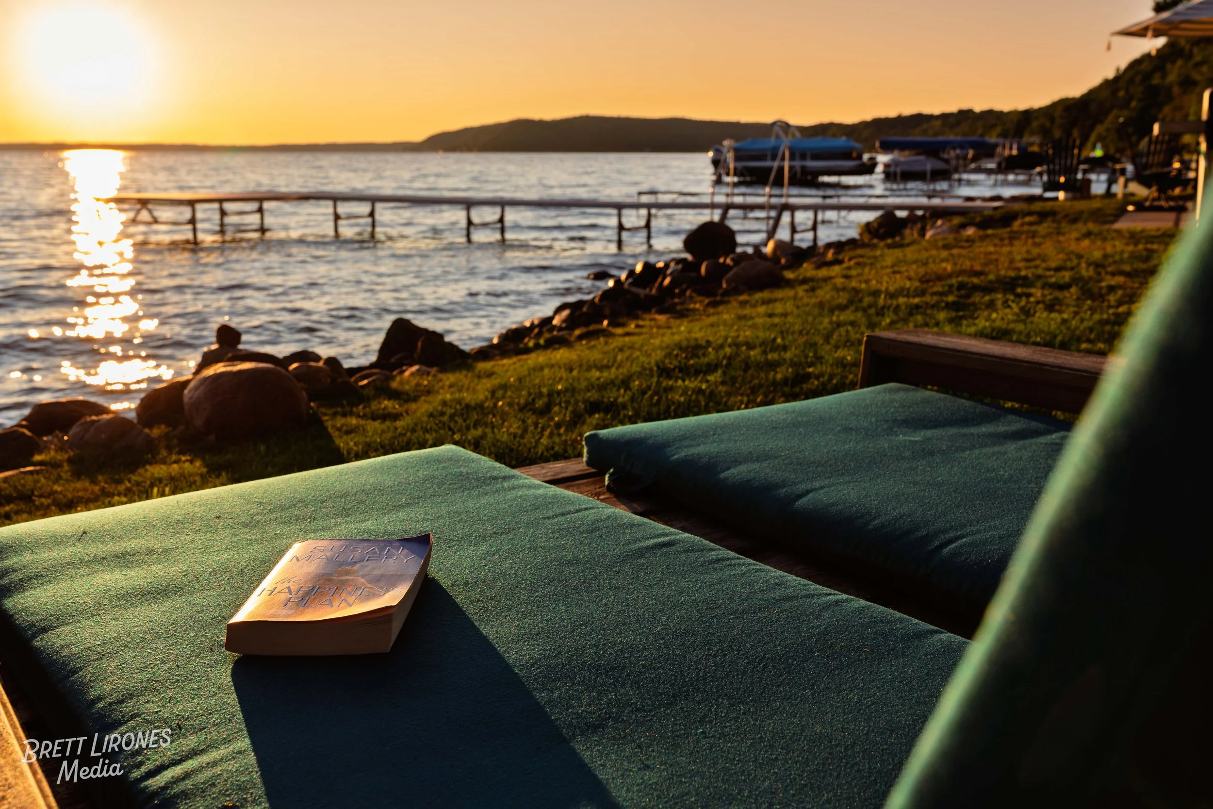 A book resting on a green cushioned chair by a lakeshore at sunset, with rocks, grass, and boats in the background.