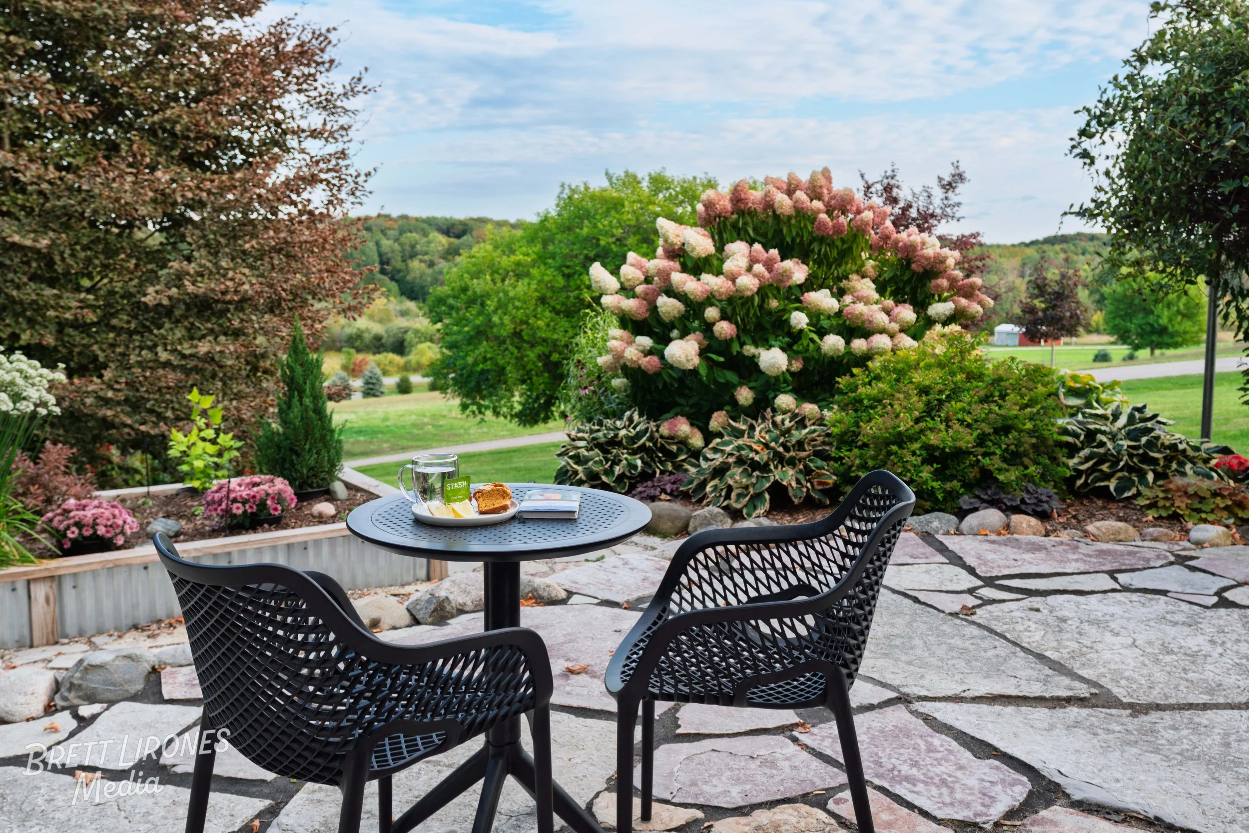 A patio with a round table and two black lattice chairs, set with a glass mug, a plate with snacks, and a book. Surrounding the patio are flower beds with pink and purple flowers, green bushes, and trees, with an open grassy field and hills in the ba