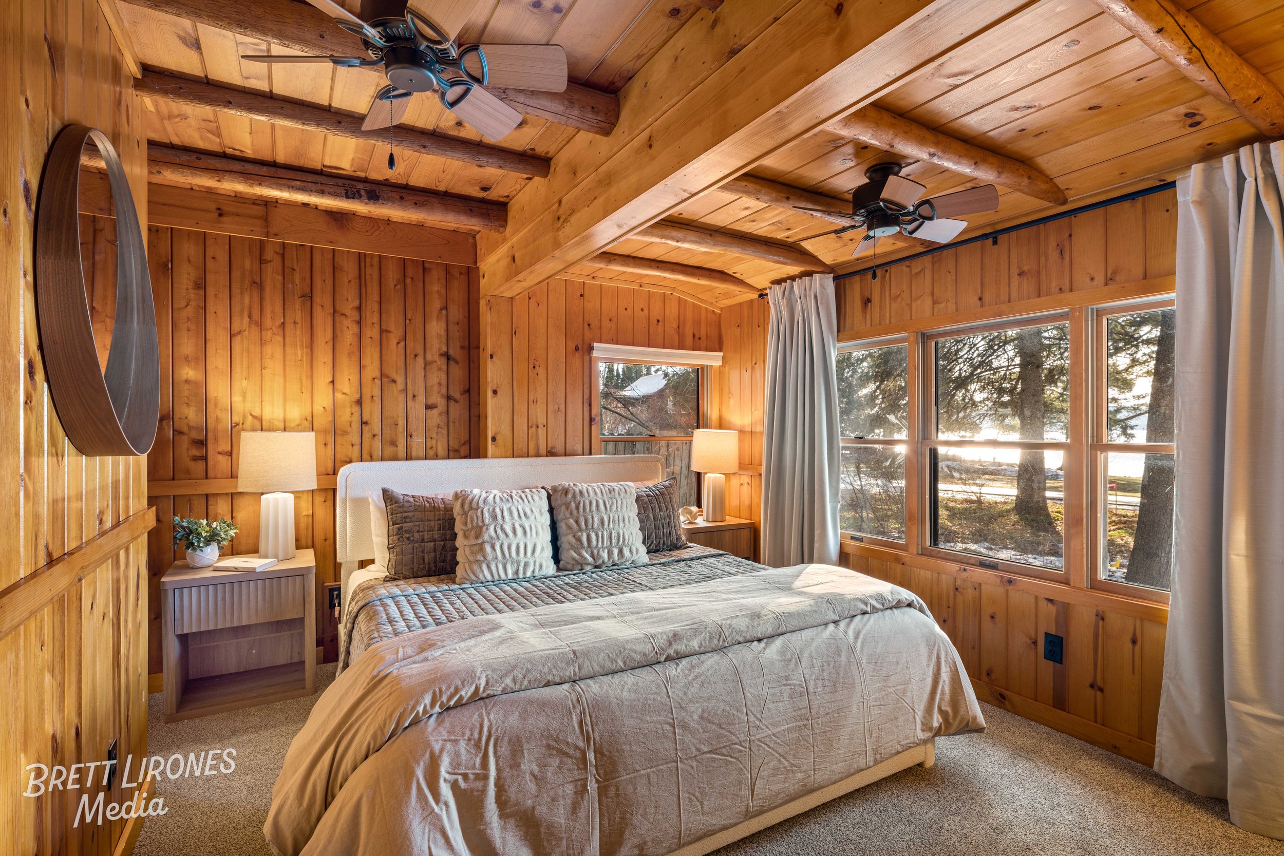 Cozy bedroom with wooden walls, large window showing trees outside, bed with gray and beige bedding, multiple pillows, nightstands with lamps, ceiling fans, and curtains.