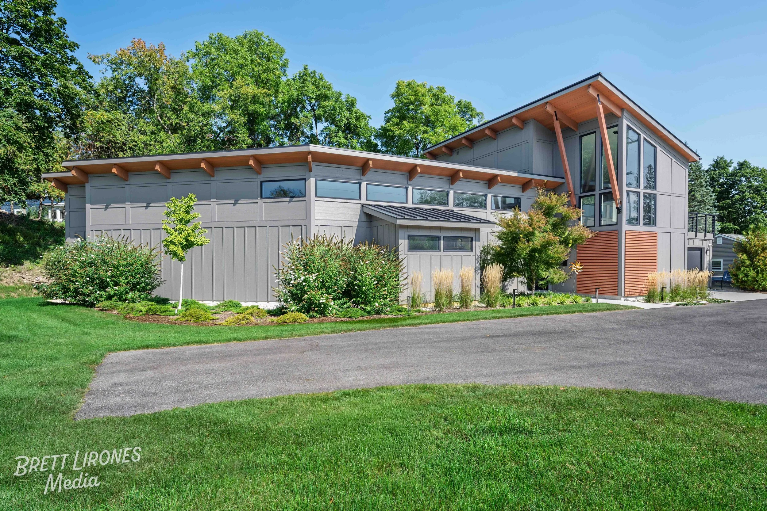 Modern gray house with large windows, wooden accents, surrounded by green lawn and trees under a clear blue sky.