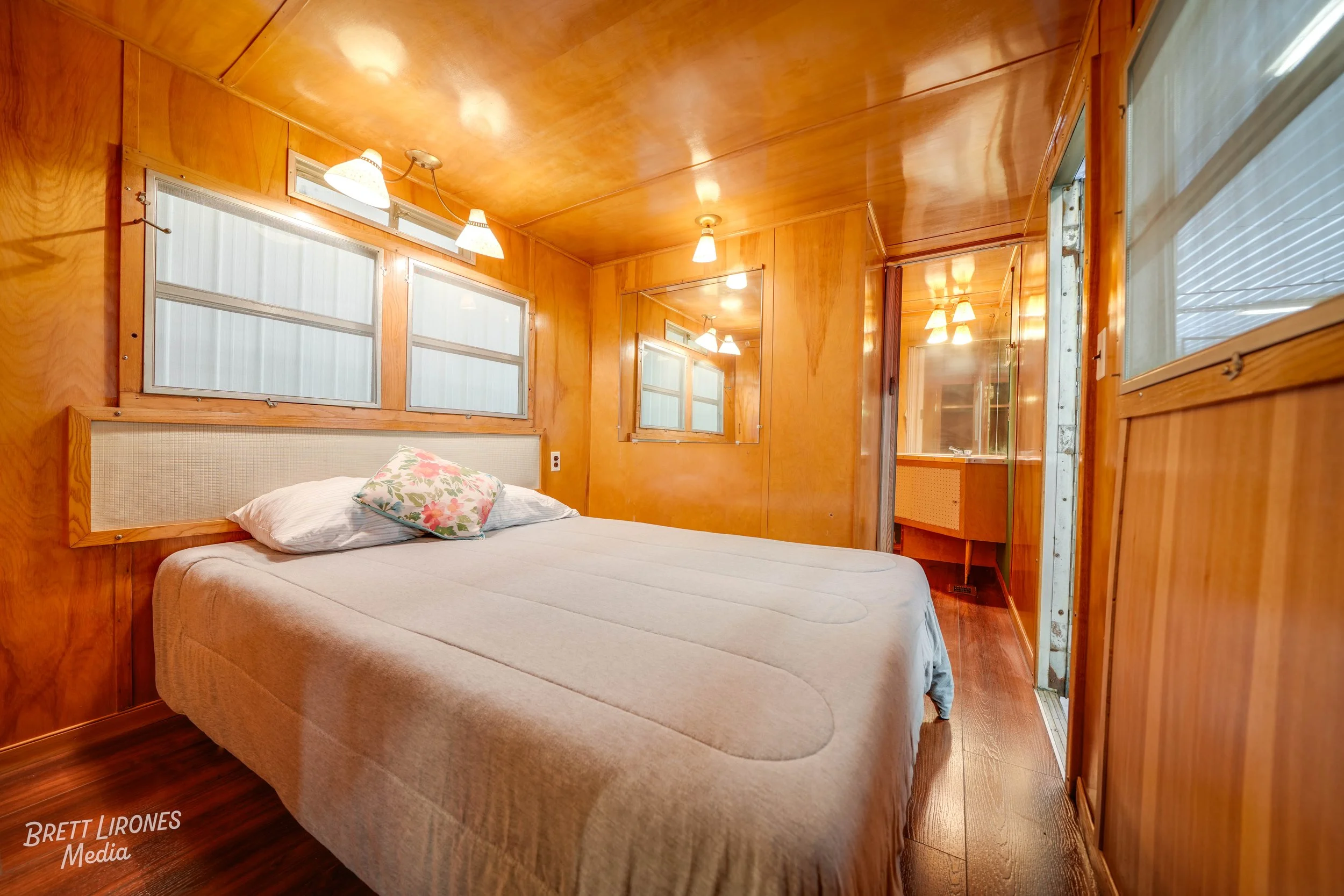 Small wooden-paneled bedroom with a bed, floral pillow, mirrored wall, and a bathroom in the adjoining space.
