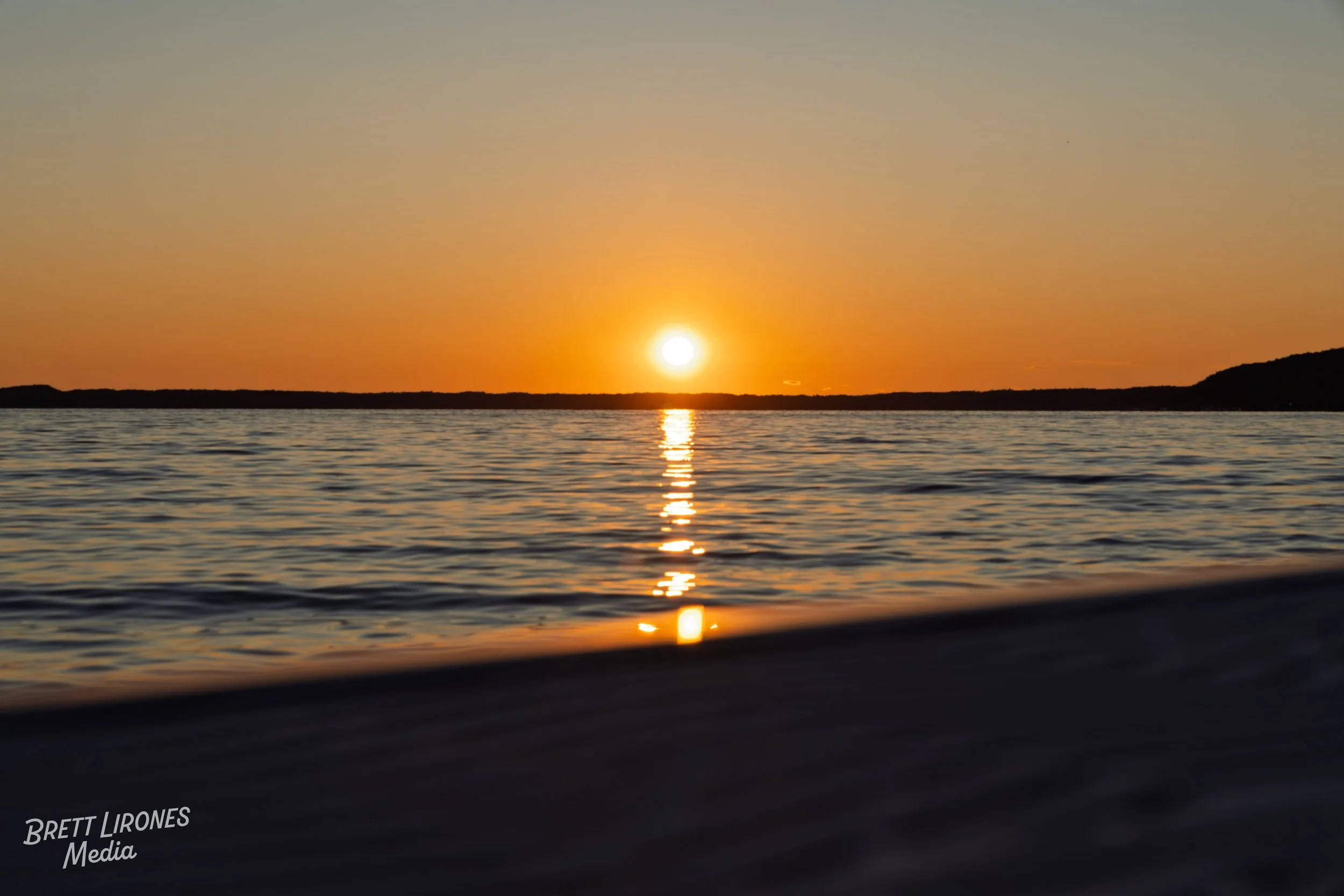 The image shows a sunset over calm water with the sun near the horizon, casting a reflection on the surface. The sky transitions from orange near the horizon to a light blue at the top. There are dark landmasses on either side of the horizon.